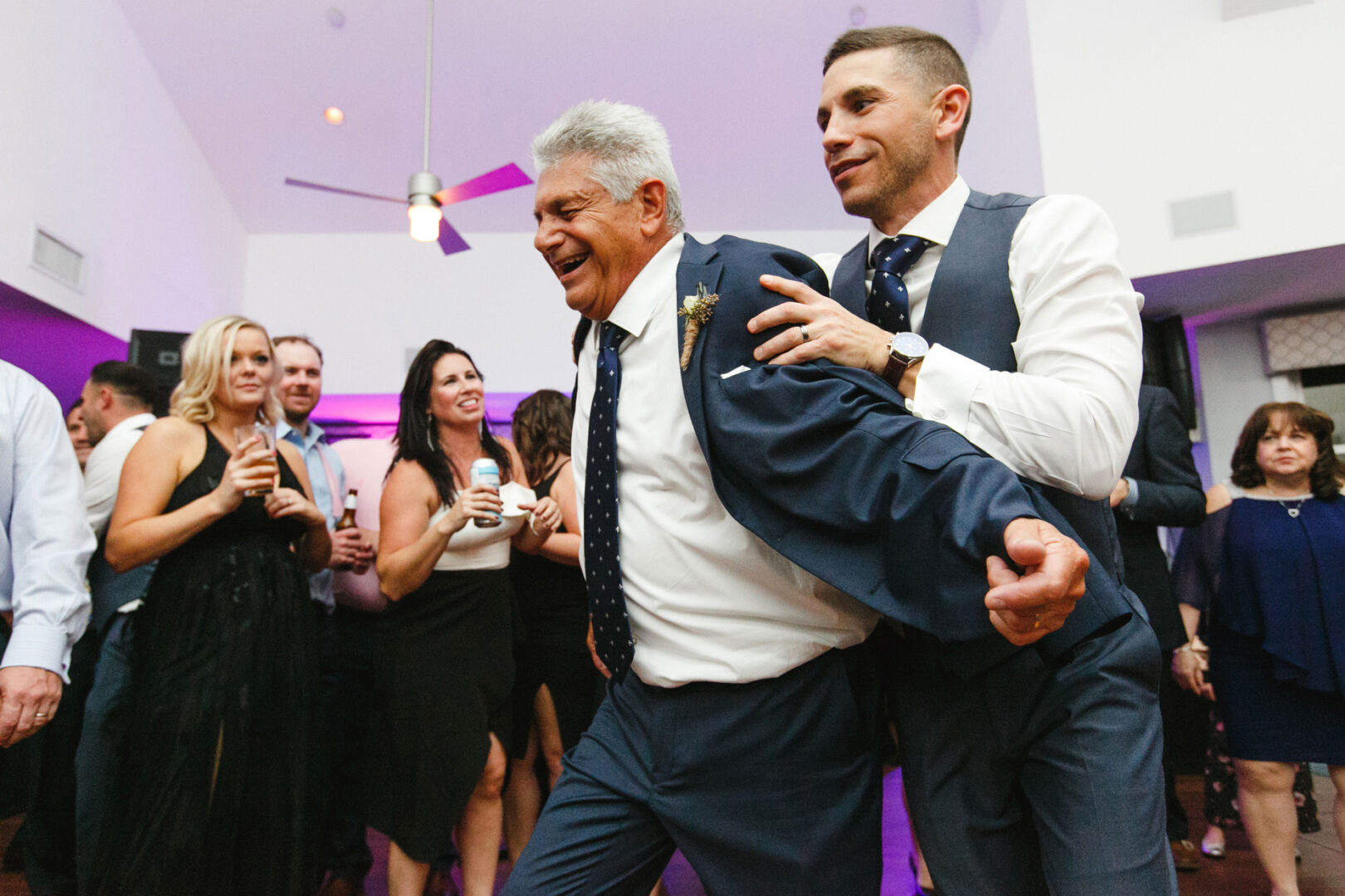 At a lively Yacht Club of Sea Isle City wedding, two men in suits dance energetically while a group of onlookers smile and cheer in the background.
