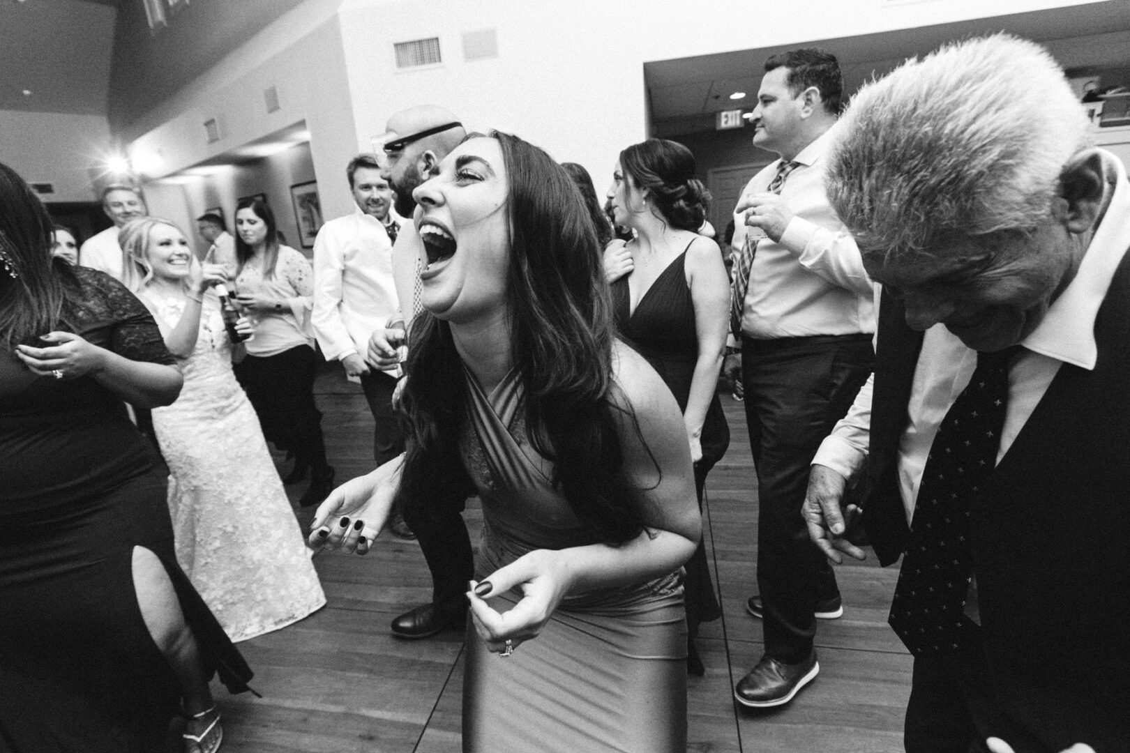 A group of people in formal attire joyfully dances and laughs at a lively indoor event, reminiscent of a spirited Yacht Club of Sea Isle City wedding.