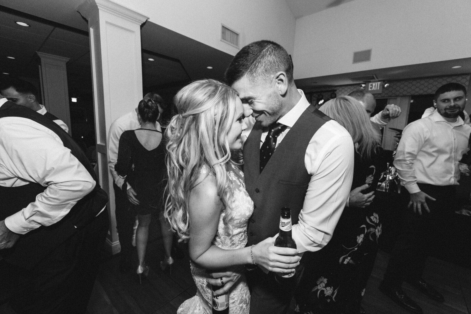 A couple in formal attire embraces and smiles at each other, drinks in hand, while enjoying a lively Yacht Club of Sea Isle City wedding. Other guests are visible in the background. Black and white photo.