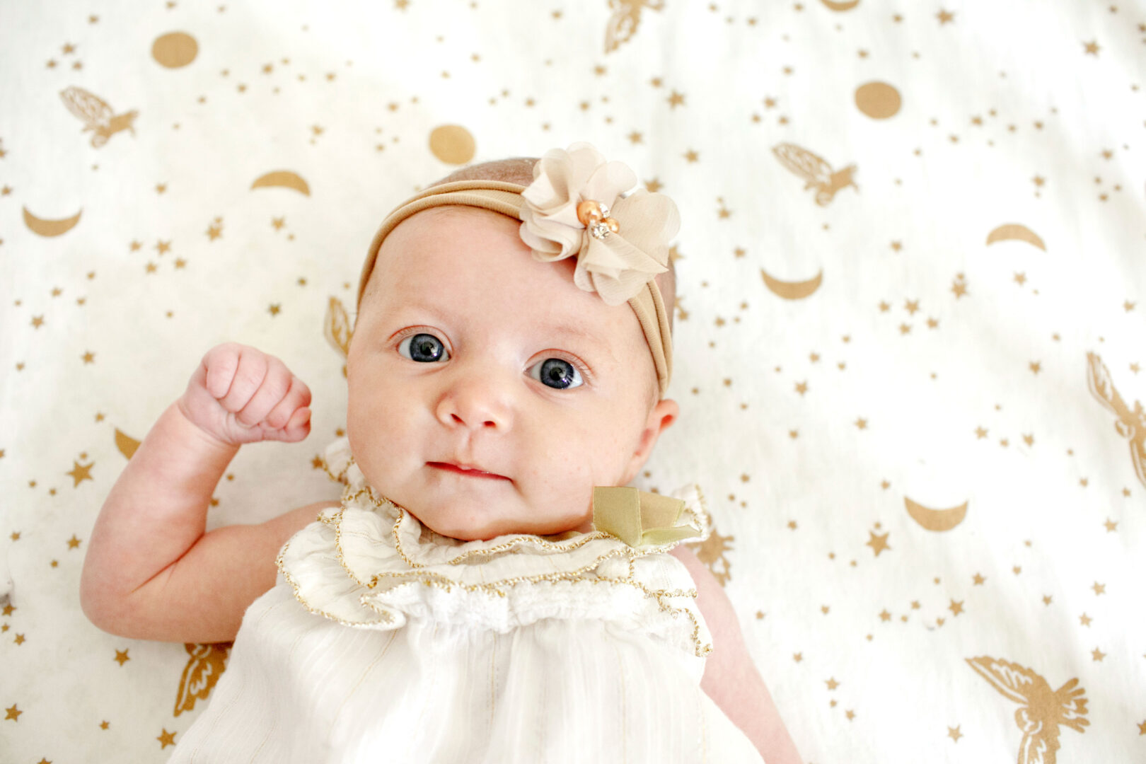 A baby wearing a beige headband adorned with a flower lies on a white blanket sprinkled with gold stars and butterflies, gazing charmingly at the camera—a picture-perfect moment reminiscent of the best Philadelphia family photos.