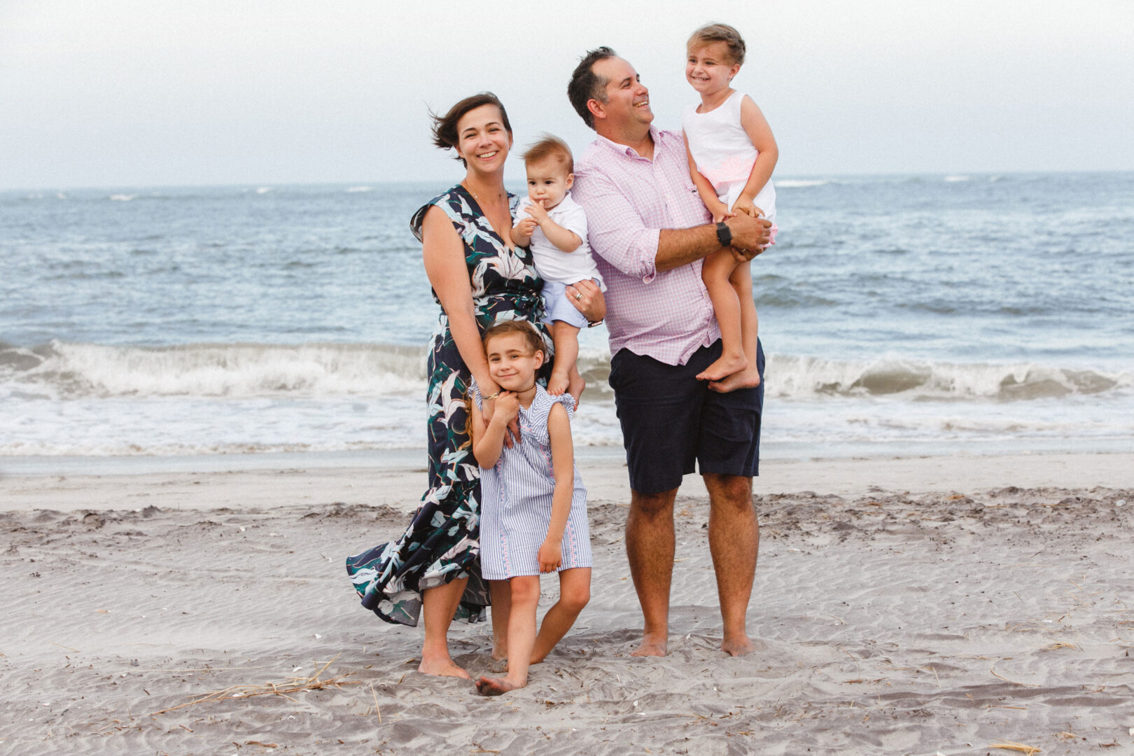 A family of five stands on a sandy beach with the ocean in the background, capturing one of their best Philadelphia family photos. The parents smile, holding two children, while another child stands in front, all dressed in casual summer clothing.