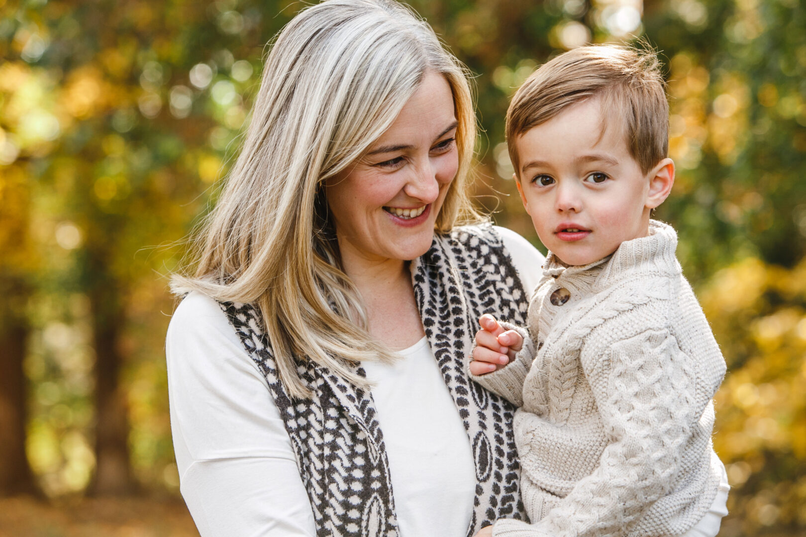 Amid the vibrant hues of autumn foliage, a woman with long hair warmly embraces a young boy in a knitted sweater. Both are smiling gently, capturing a moment that would surely be among the best Philadelphia family photos.
