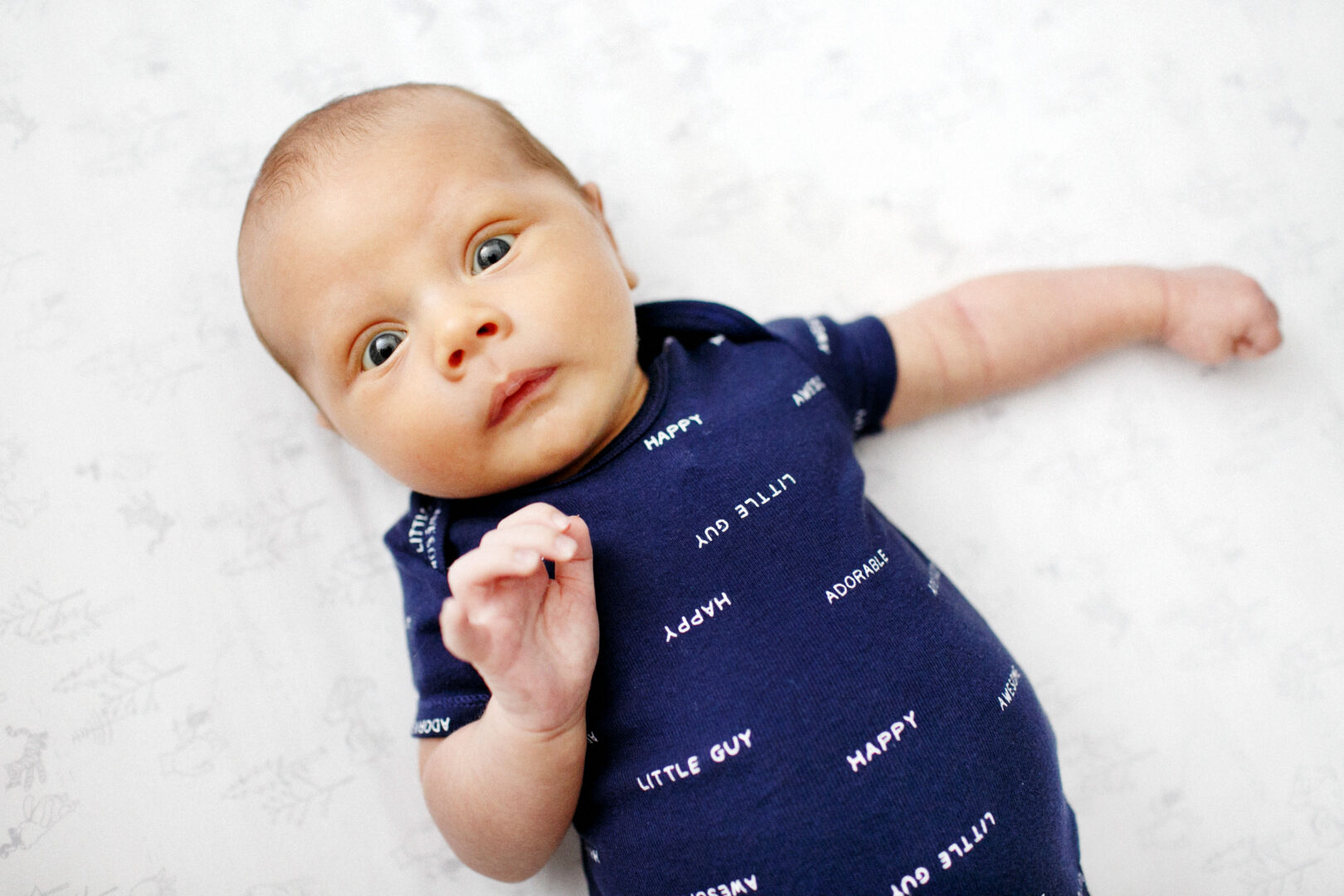 Baby lying on a white surface, wearing a dark blue onesie with text that reads "HAPPY" and "LITTLE GUY," capturing the essence of the best Philadelphia family photos.
