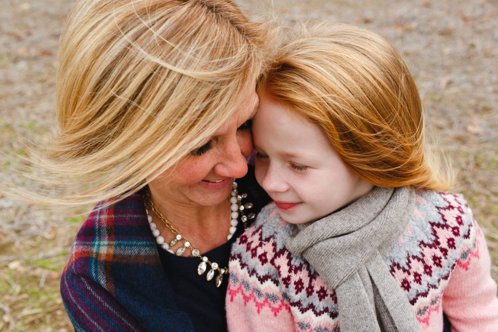 A woman and a young girl with red hair share a warm embrace outdoors. The woman wears a colorful shawl and necklace, while the girl sports a patterned sweater and scarf. This captures one of the best Philadelphia family photos, showcasing their genuine joy and connection.