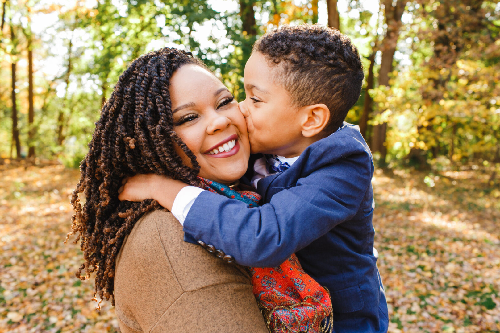 In one of the best Philadelphia family photos, a child in a blue suit kisses a smiling woman with braided hair in a brown coat. They stand nestled in a wooded area, surrounded by the warm hues of autumn leaves carpeting the ground.