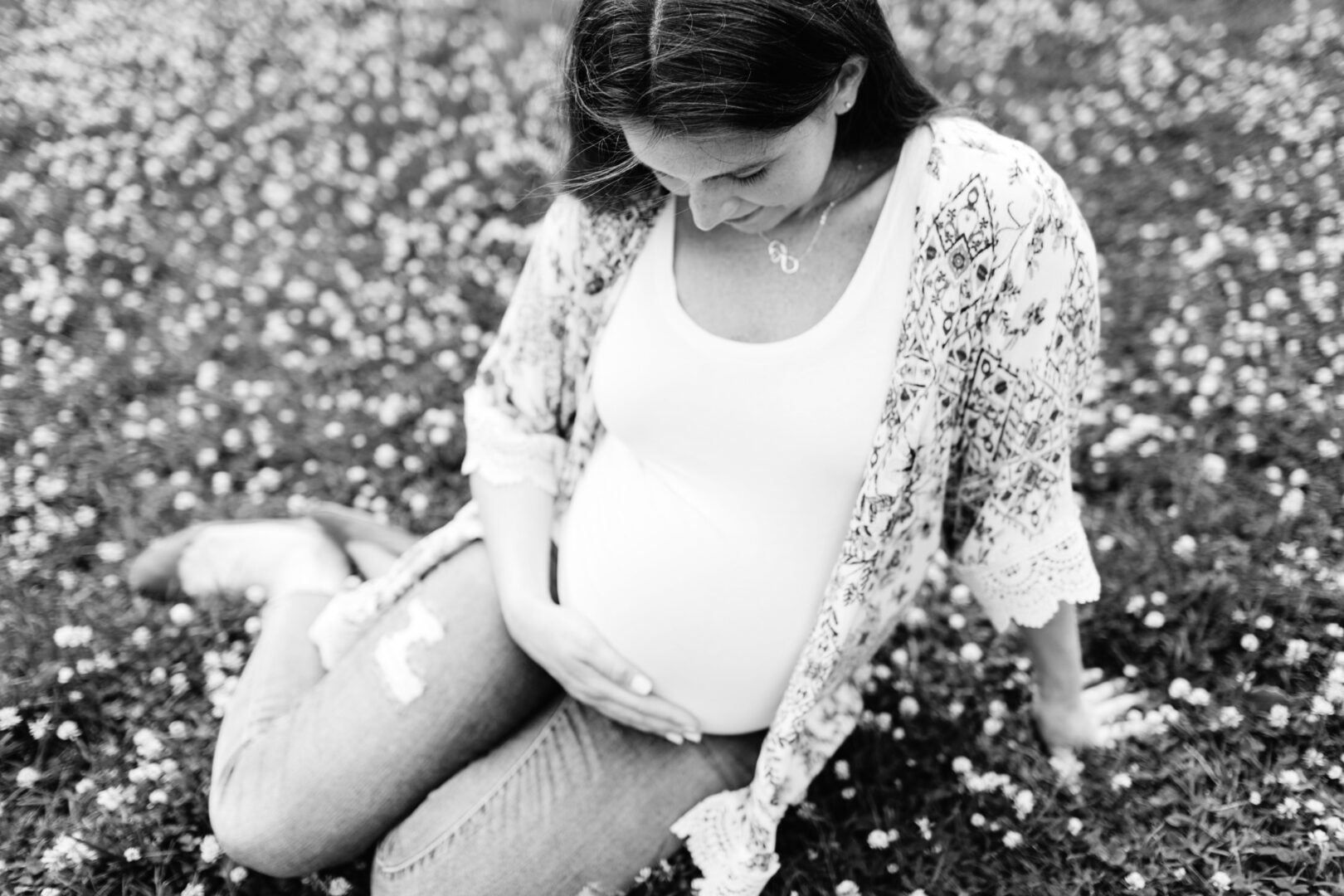 In this timeless black and white image, a serene pregnant woman sits gracefully on grass dotted with small flowers. She's clad in a floral cardigan, white top, and jeans, gently touching her belly and gazing downward—a perfect capture for Best Philadelphia Family Photos.