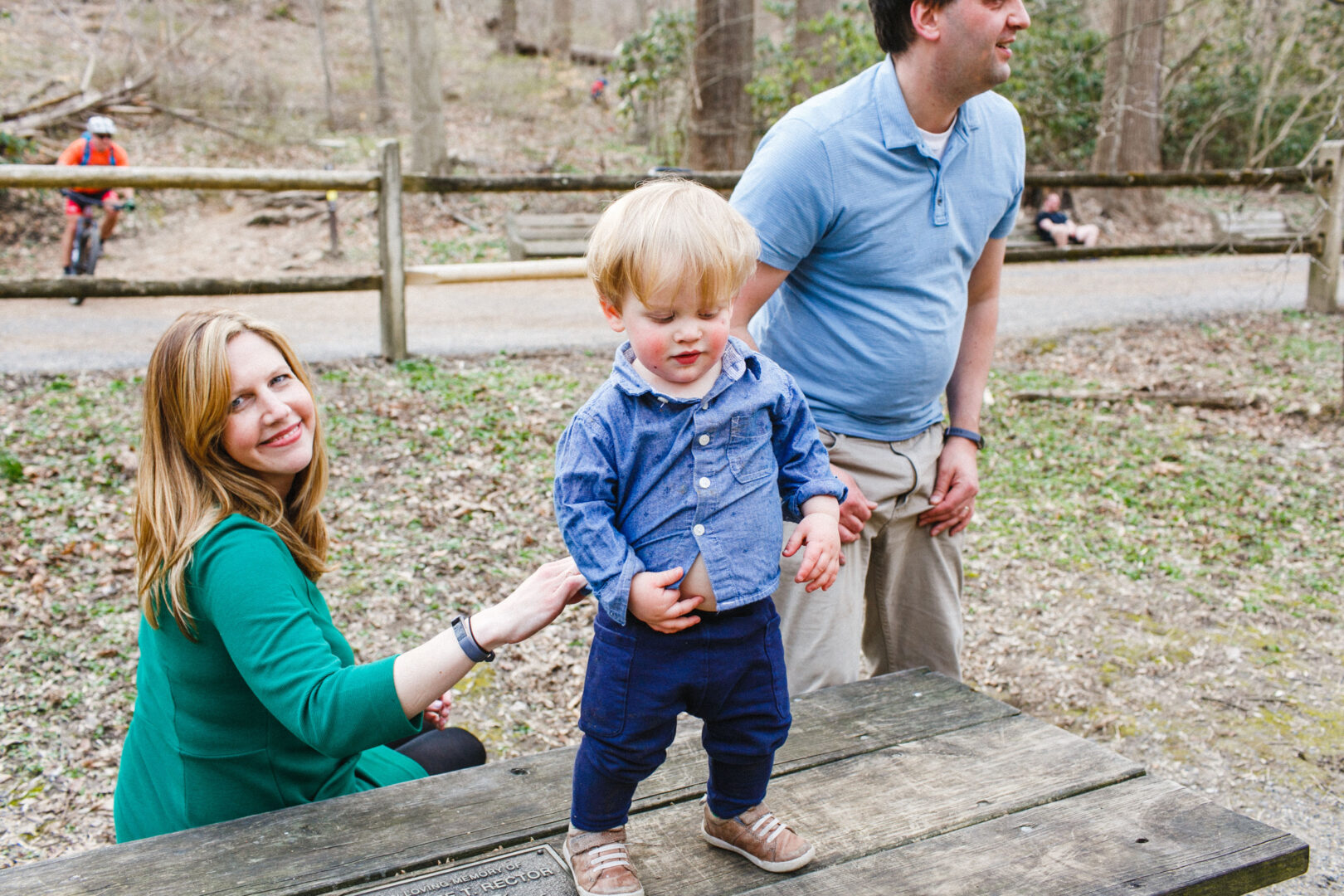 A child stands on a wooden table outdoors, capturing the essence of Best Philadelphia Family Photos, with a woman nearby and a man slightly behind. Trees and a pathway create the perfect backdrop for this memorable scene.