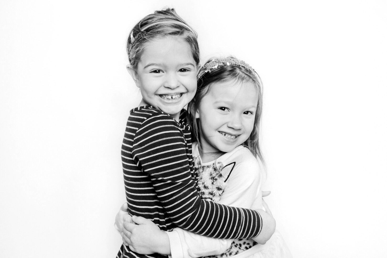 Two smiling children hug each other, both wearing casual clothes. One is in a striped shirt, the other in a patterned top. Capturing this heartfelt moment against a plain white backdrop makes for one of the best Philadelphia family photos.