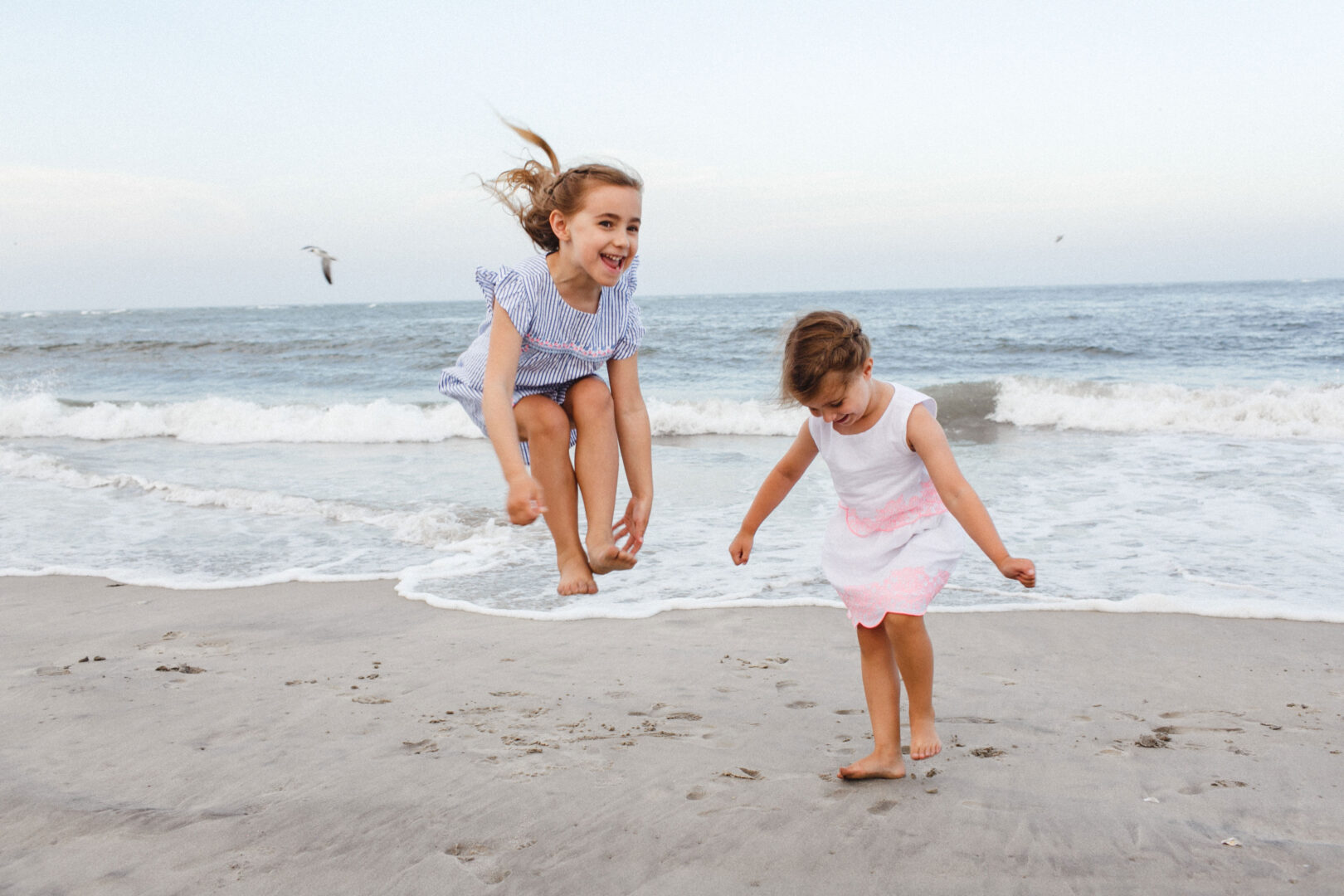 Two girls play on a sandy beach, one jumping in the air while the other walks. The ocean stretches behind them, dotted with seagulls. It's a scene reminiscent of the best Philadelphia family photos, capturing carefree childhood moments by the sea.