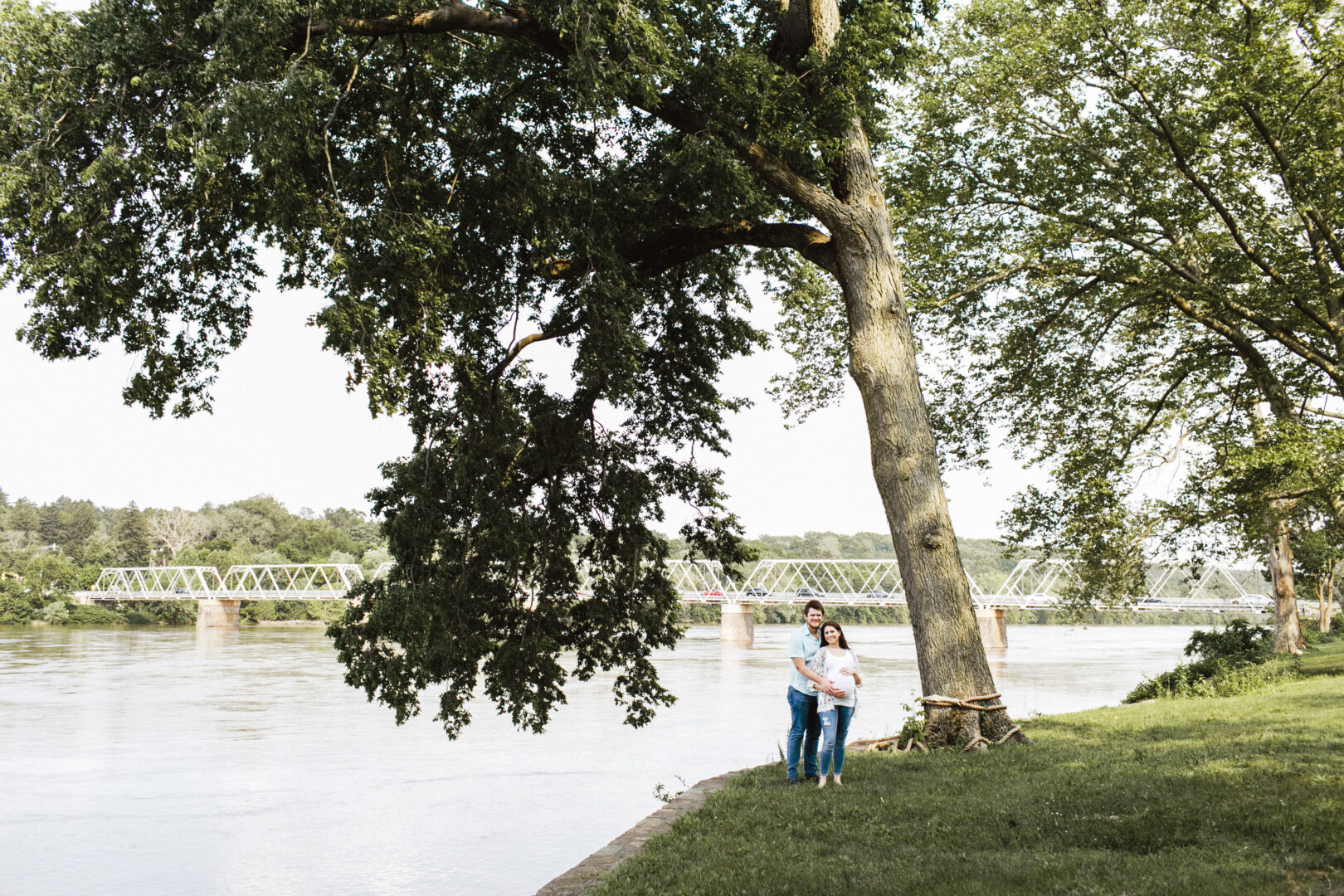 A couple stands by a large tree near a river, with a bridge in the background and lush greenery surrounding them, capturing one of the best Philadelphia family photos.