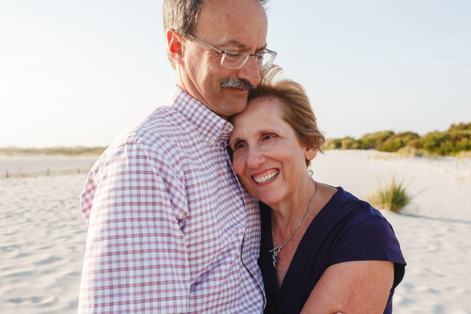 A couple embraces on a sandy beach, with the woman smiling and leaning on the man's chest. He wears glasses and a checkered shirt. The scene evokes the warmth of best Philadelphia family photos, set against a clear sky and distant vegetation.