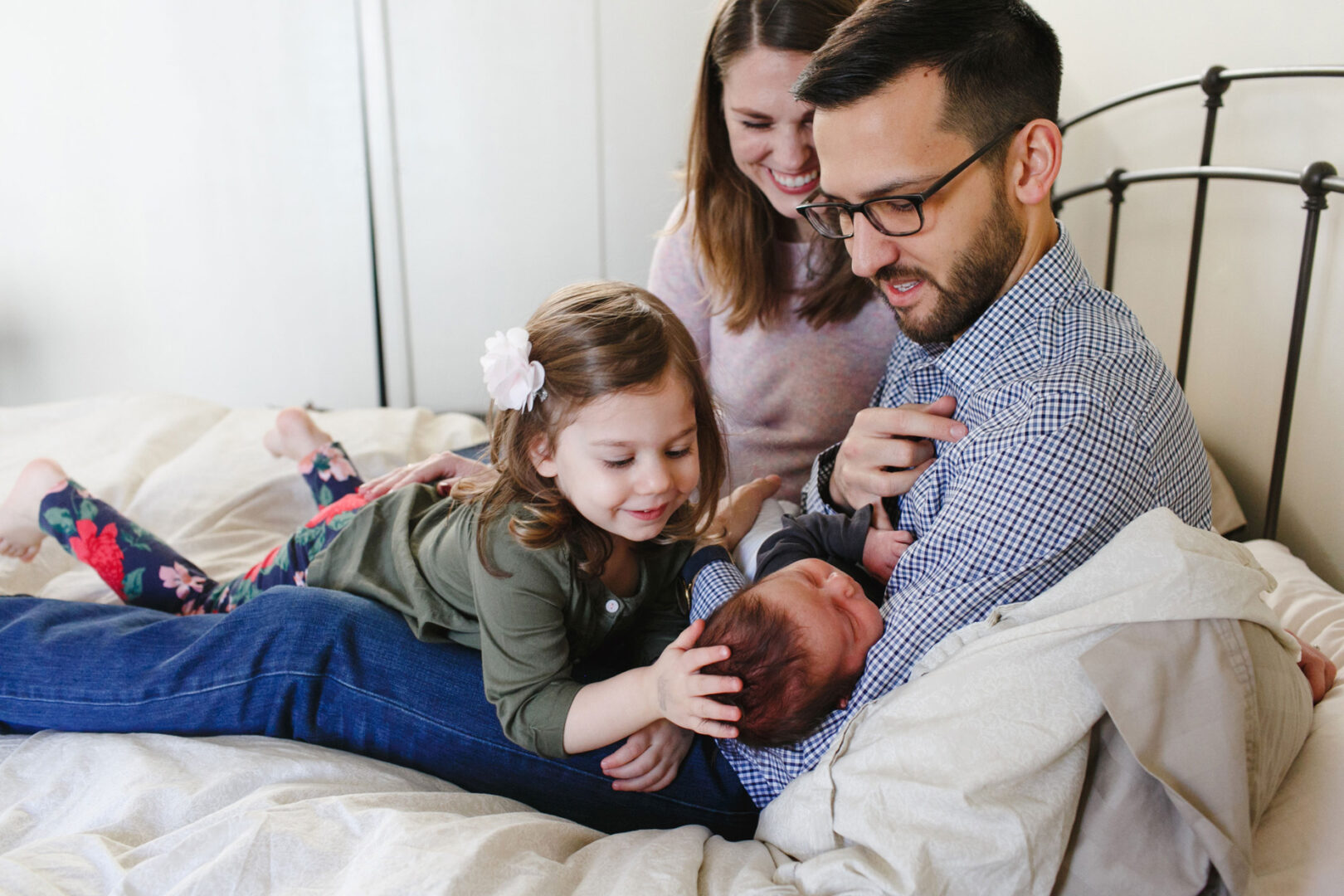 A family on a bed; an adult man holds a newborn during their Newborn Session in Philadelphia. A young girl reaches towards the baby, and an adult woman smiles in the background.