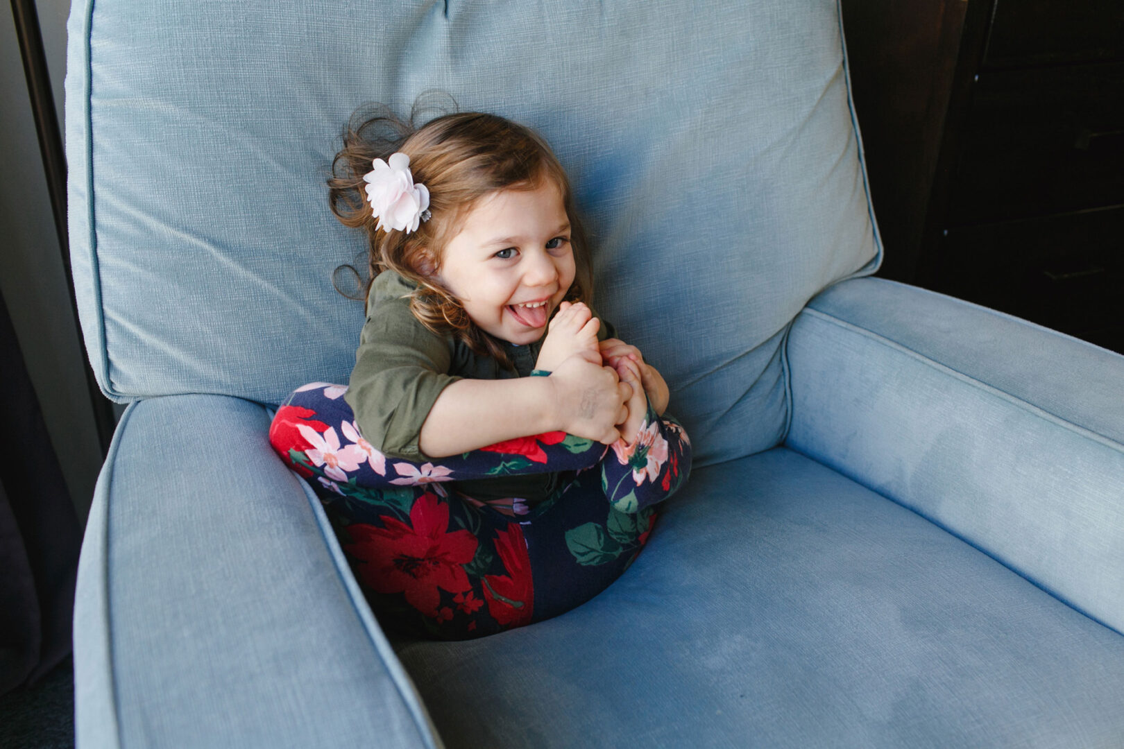 During a vibrant newborn session in Philadelphia, a young girl is sitting on a blue chair, playfully holding her foot. She's wearing charming floral pants and a cute hair accessory, adding to the delightful atmosphere of the photo shoot.