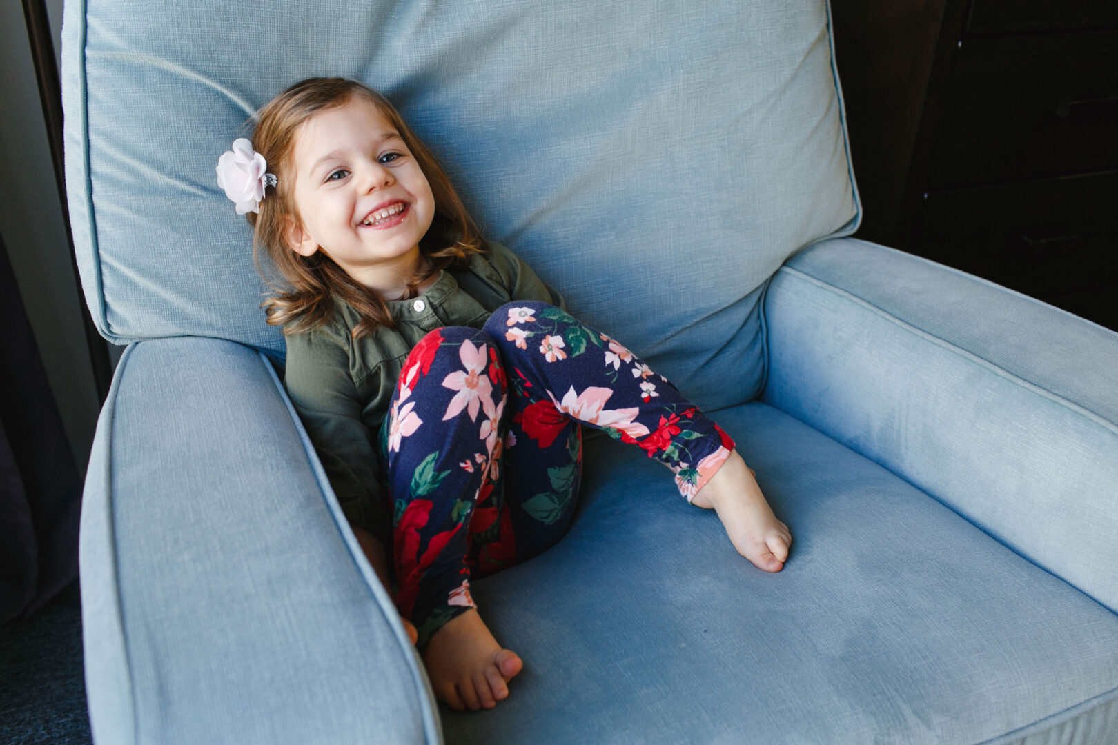 A young girl with brown hair adorned with a white bow sits smiling in a large blue armchair, wearing a green shirt and floral pants, reminiscent of the comfort and warmth found in a Newborn Session in Philadelphia.