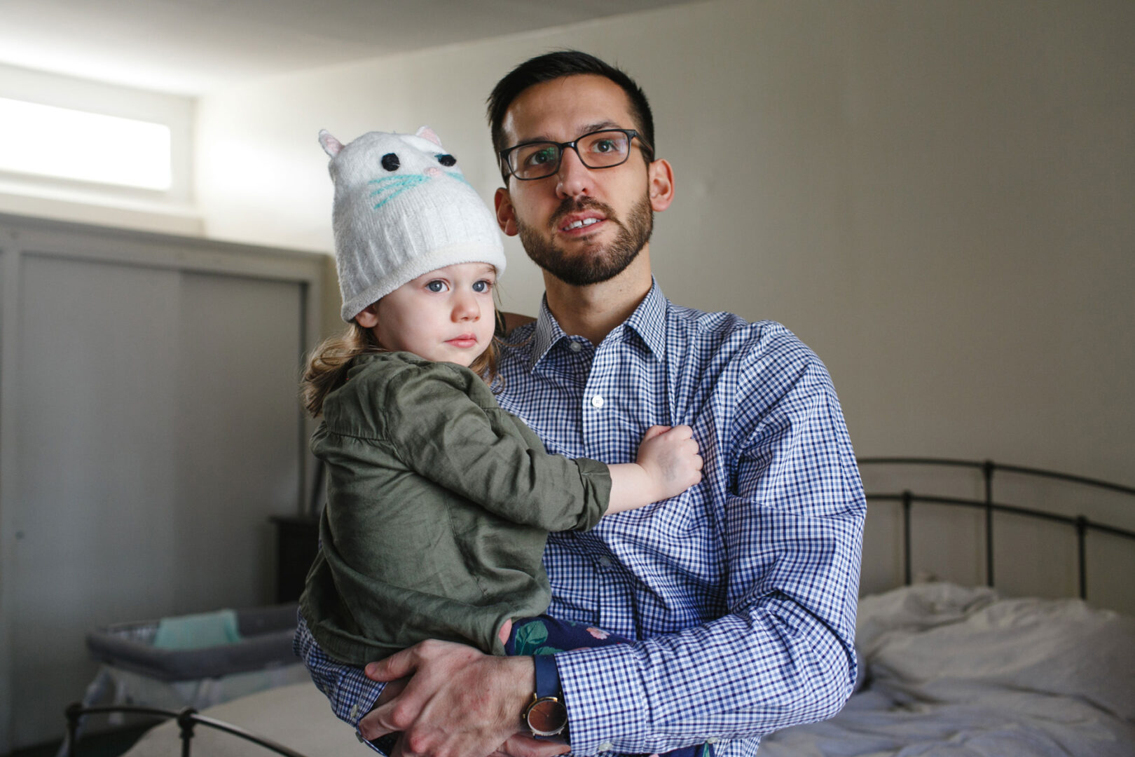 A man cradles a child, the little one snug in a whimsical white beanie with ears and a face design. In the background, a bed and closed closet doors hint at the serene setting of their newborn session in Philadelphia.