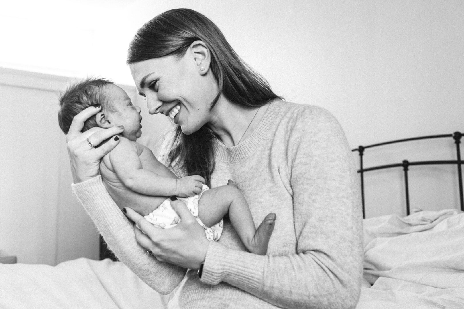 In a cozy bedroom setting, a woman smiles tenderly while holding a baby close, their noses touching. This black and white photo captures the warmth of a Newborn Session in Philadelphia.