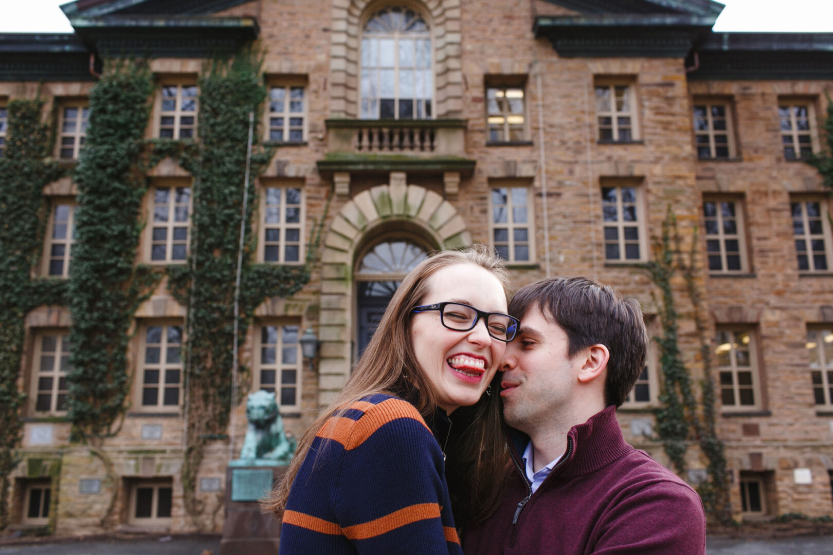 A woman and a man stand closely in front of a historic building with ivy-covered walls, capturing the charm of their Princeton engagement session. The woman is smiling as the man leans towards her, enveloping them both in a moment that feels timeless against the storied backdrop.