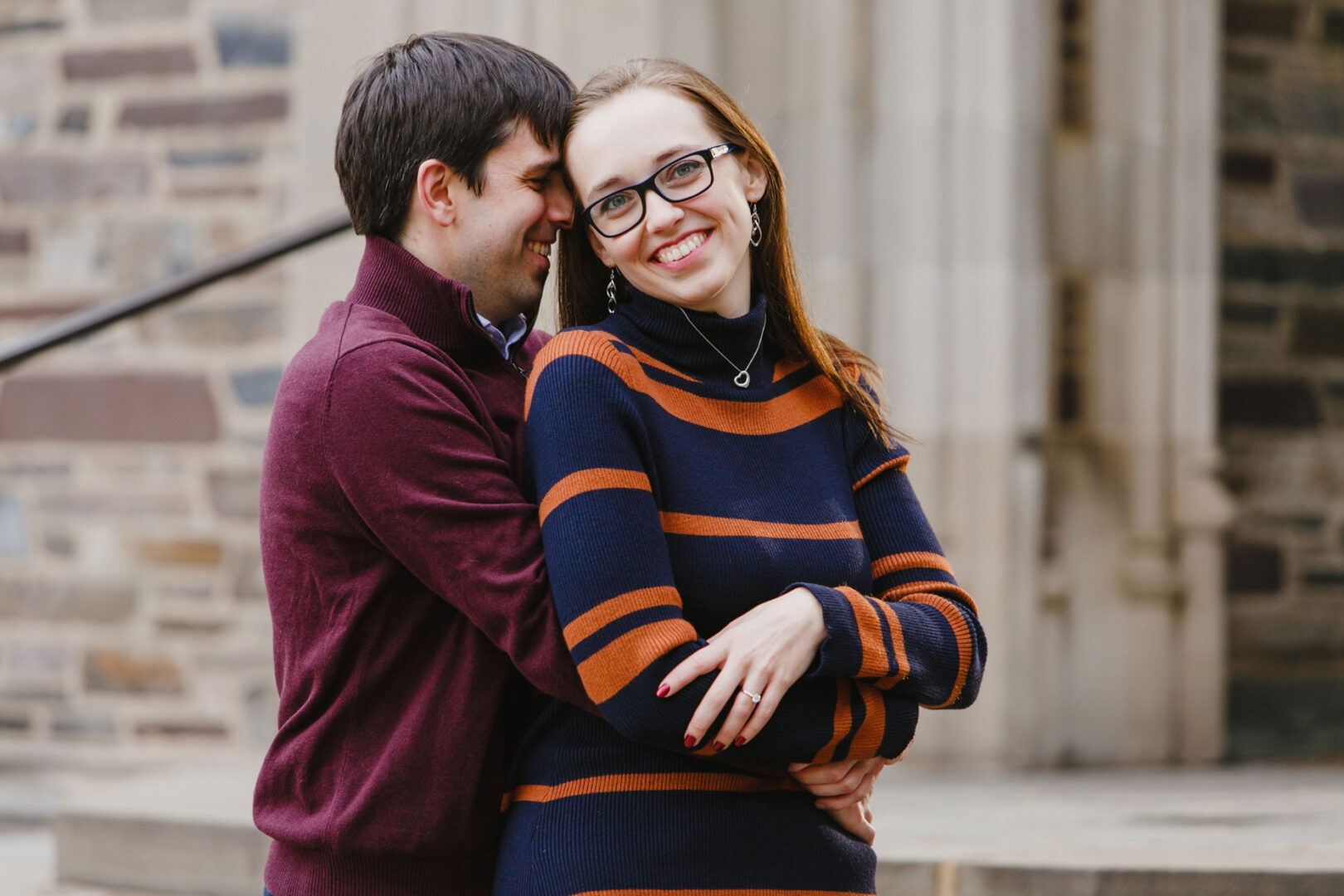 A man in a maroon sweater hugs a smiling woman in a navy and orange striped sweater during their enchanting Princeton engagement session. They're standing lovingly in front of the timeless backdrop of a stone building.