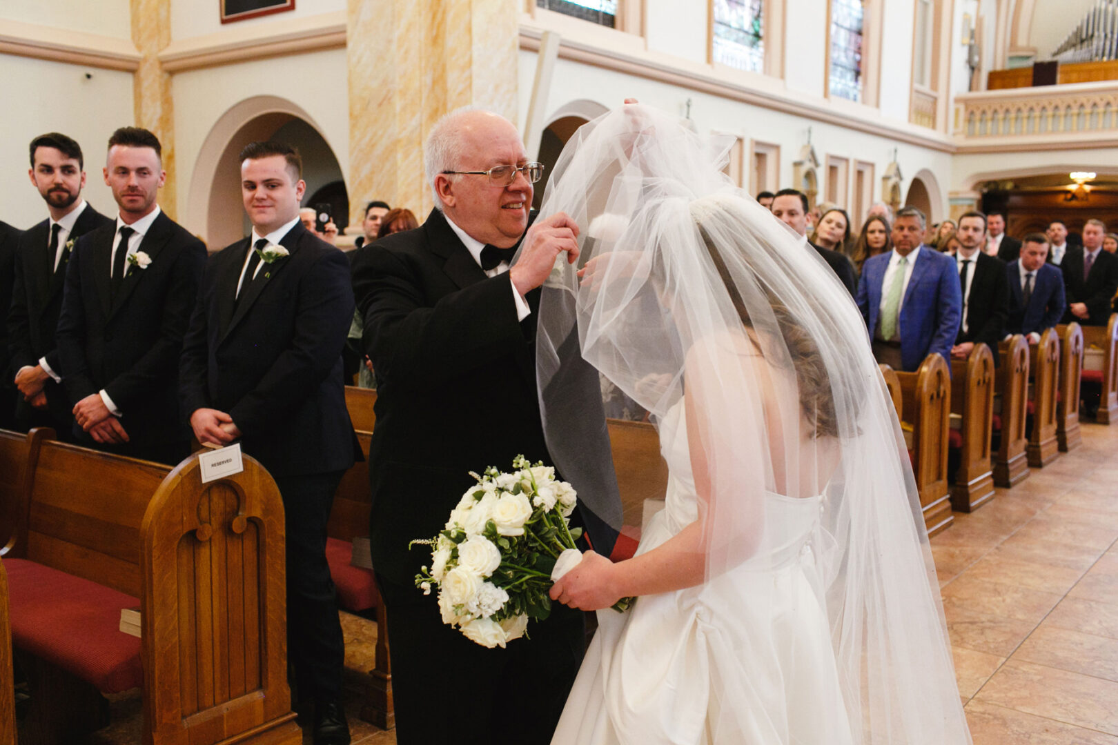 In the grand setting of Congress Hall, a man lifts the bride's veil in the church as groomsmen and guests look on. The bride clutches a bouquet of white flowers, surrounded by an aura of joy and love.