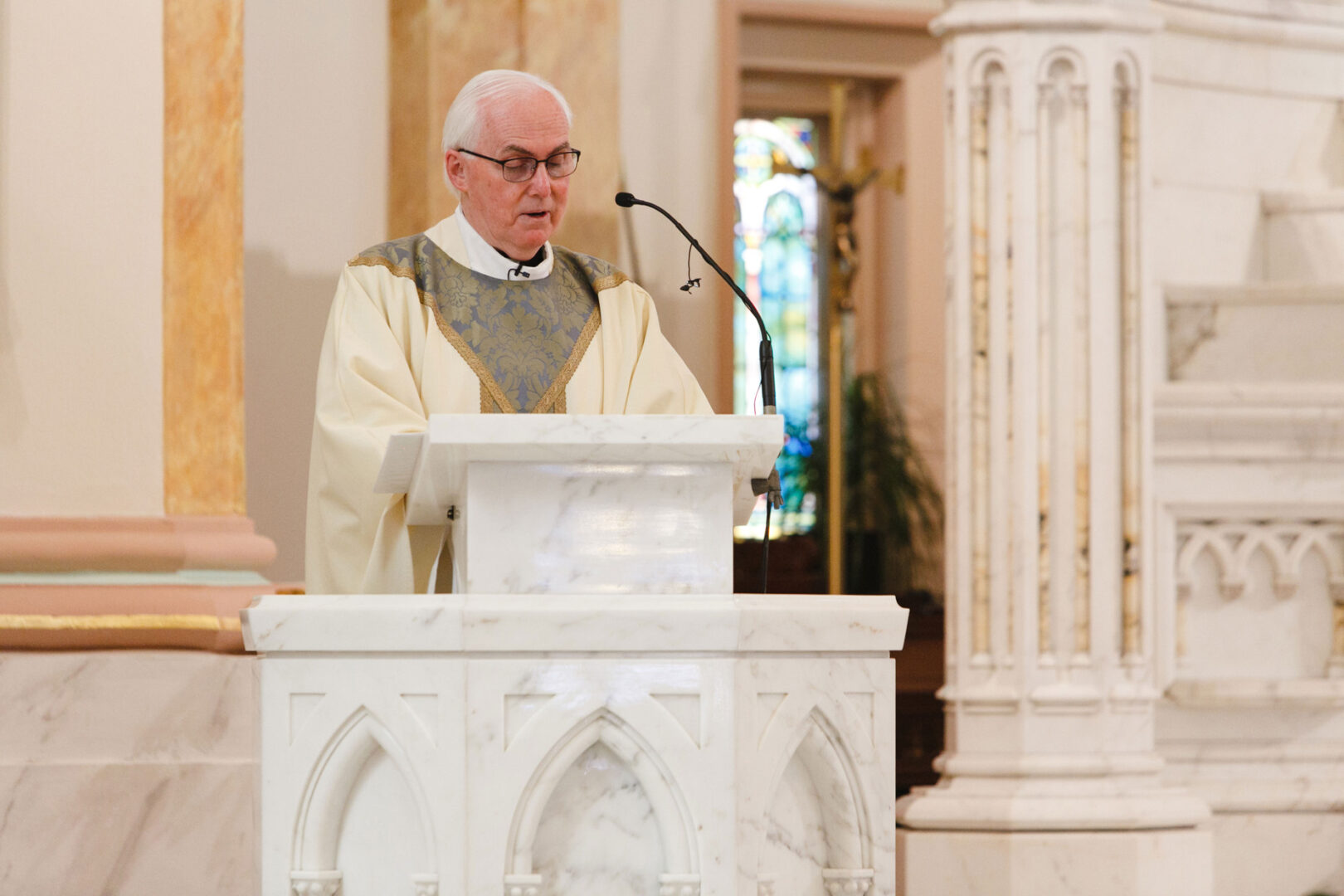 A priest in liturgical attire speaks from a white marble pulpit inside a church with stained glass windows, reminiscent of the grandeur often seen at weddings held in Congress Hall.