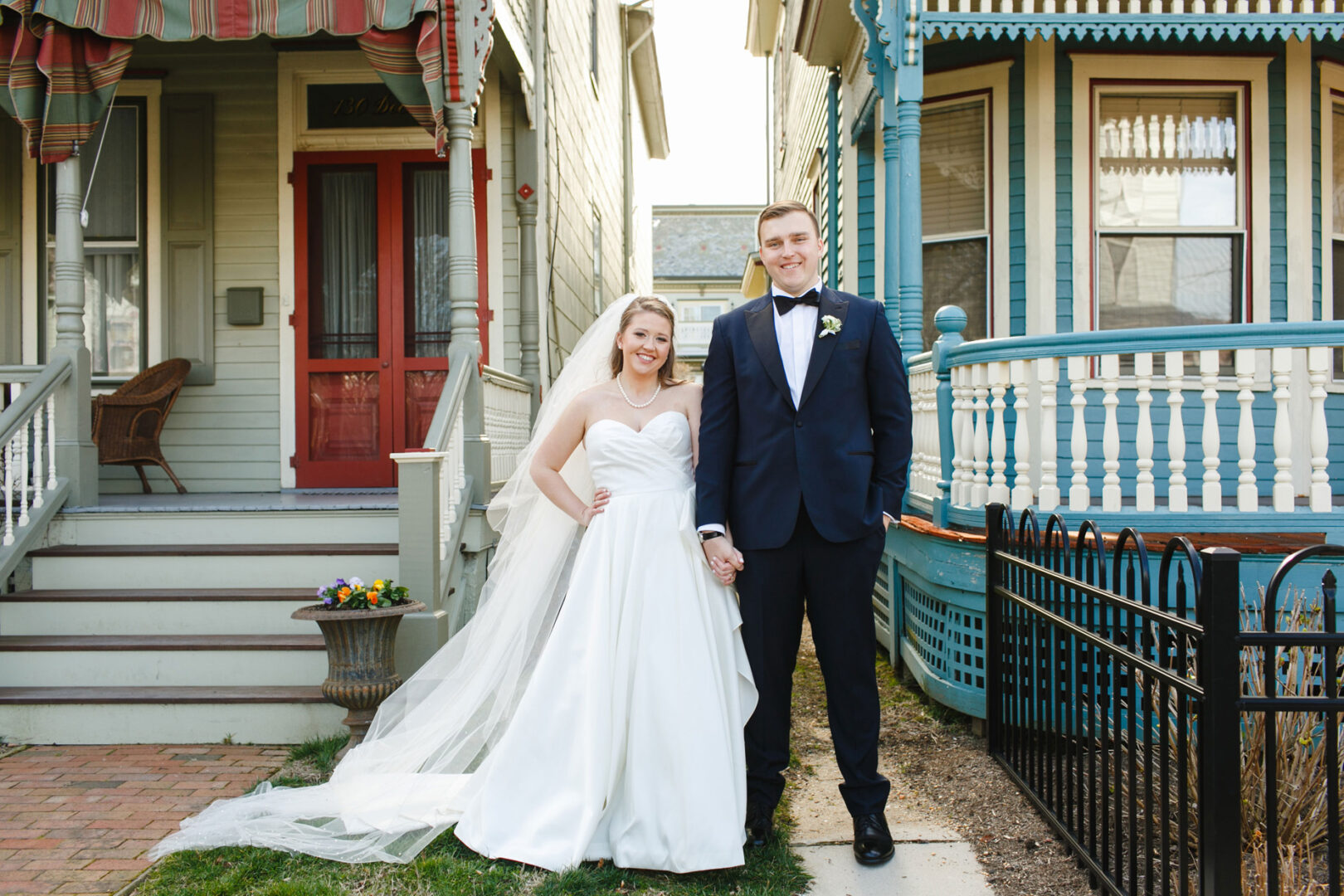 The bride in a white gown and the groom in a navy suit stand holding hands, embodying elegance, as they pose before two traditional houses with ornate porches—a scene reminiscent of a classic Congres Hall wedding.