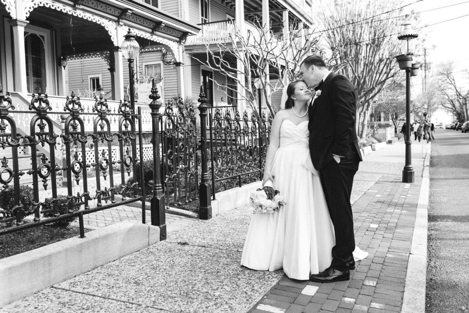 A bride and groom share a kiss on a sidewalk beside a wrought iron fence, with a large house reminiscent of Congress Hall in the background. The bride holds a bouquet.