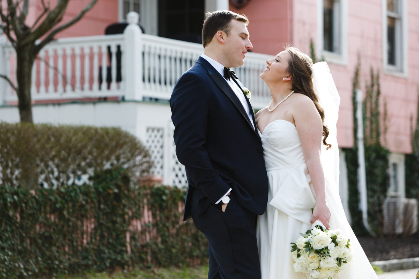 The bride and groom stand outdoors, facing each other near a pink building at Congress Hall. The bride holds a bouquet of white flowers, and both are dressed in formal attire.