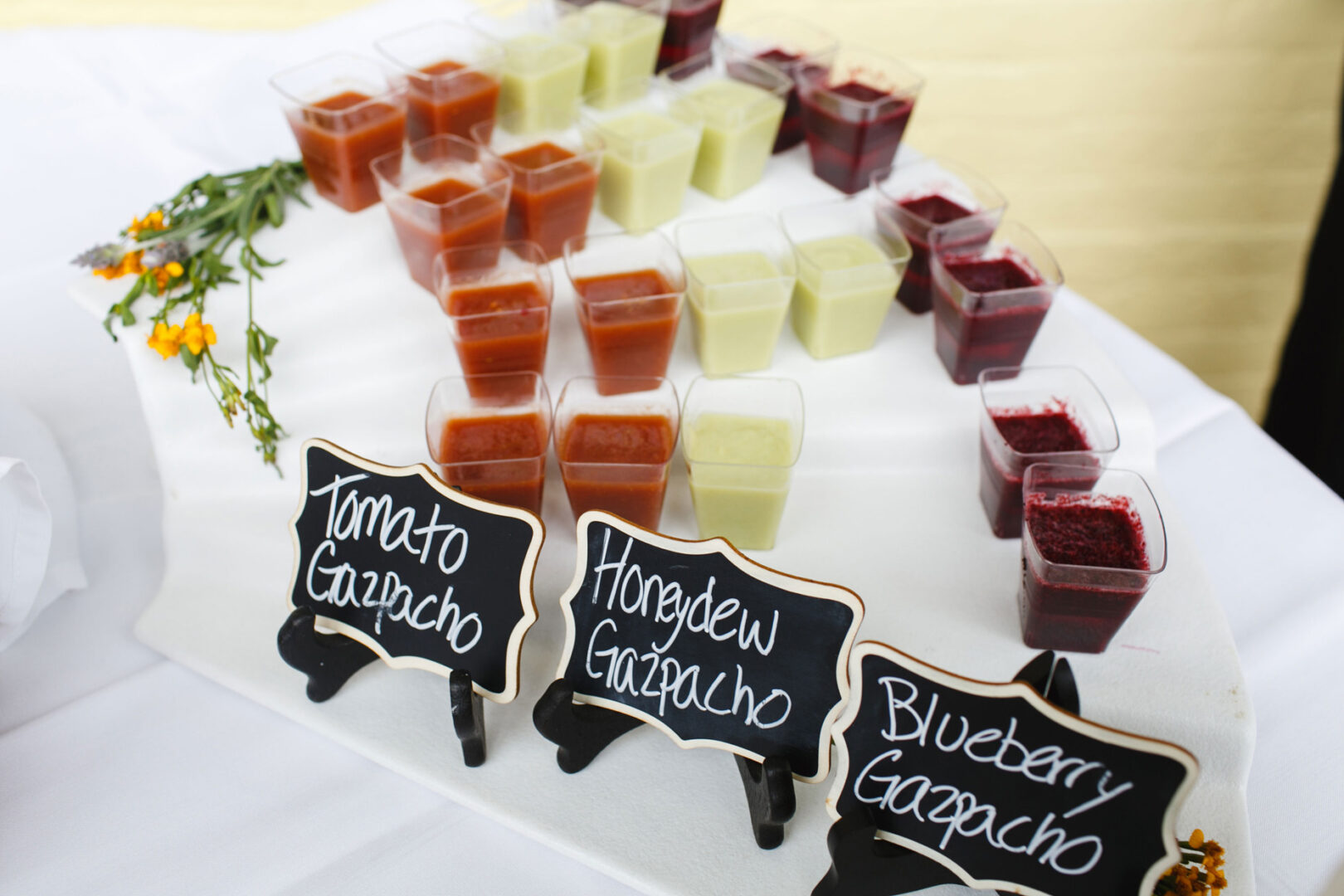 At the Congres Hall Wedding, small cups of tomato, honeydew, and blueberry gazpacho grace a white tray, accented with labeled chalkboard signs and a delicate floral garnish.