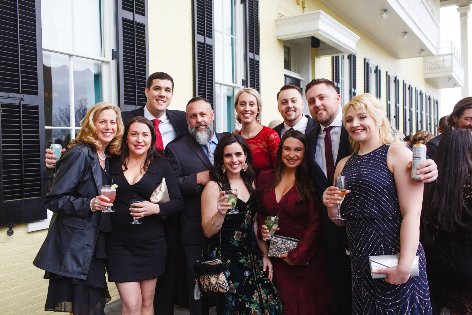 A group of people, elegantly dressed for a Congres Hall wedding, smiles warmly while holding drinks, standing outdoors in front of a building with striking black shutters.