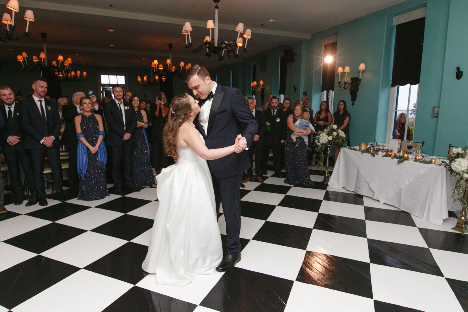 At the elegant Congres Hall wedding, a couple dances gracefully in formal attire on a checkered floor, surrounded by admiring guests.