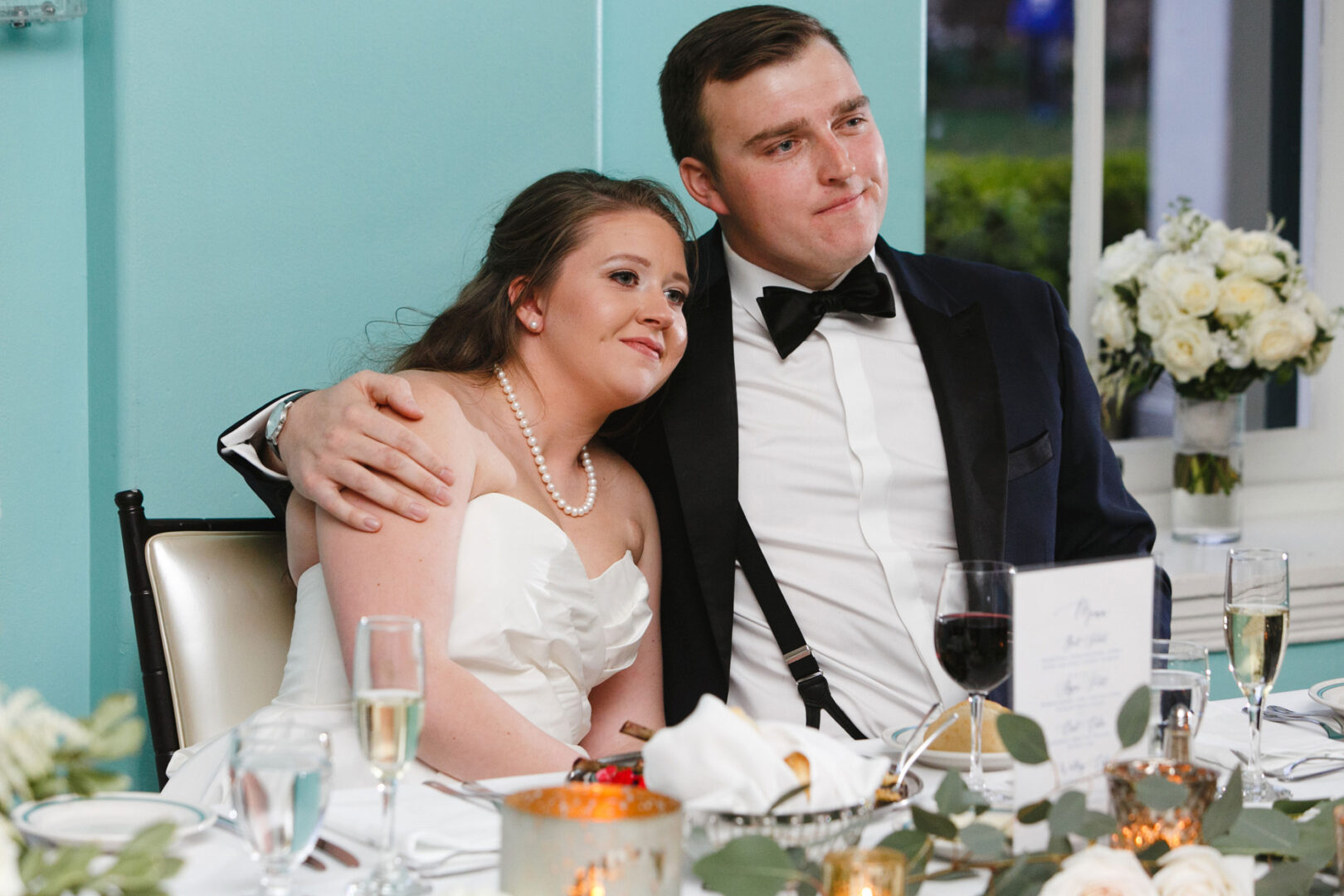 At a picturesque Congres Hall wedding, a bride in her white dress and a groom in his black tuxedo sit at a table, embracing. Surrounded by plates and glasses, their joy fills the elegant setting of this special day.