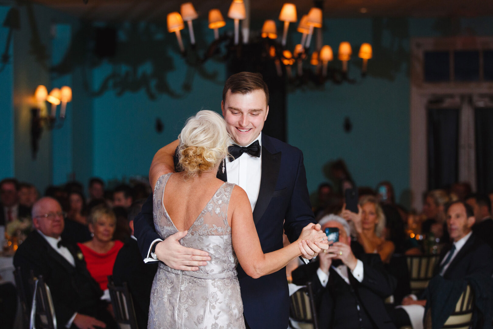A man in a tuxedo elegantly dances with a woman in a formal dress at a charming Congres Hall wedding, with seated guests enjoying the celebration in the background.