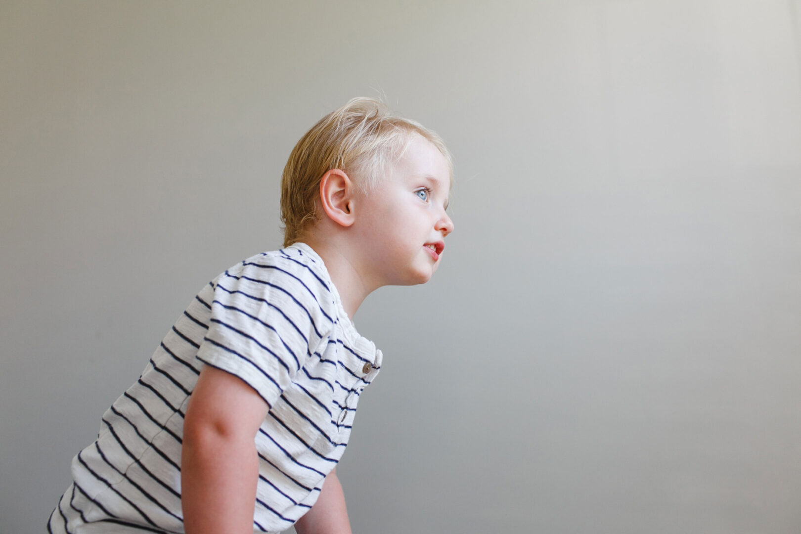 A young child with blond hair, wearing a striped shirt, looks curiously to the side against a plain background, embodying the genuine charm seen in Collegeville Family Photos.