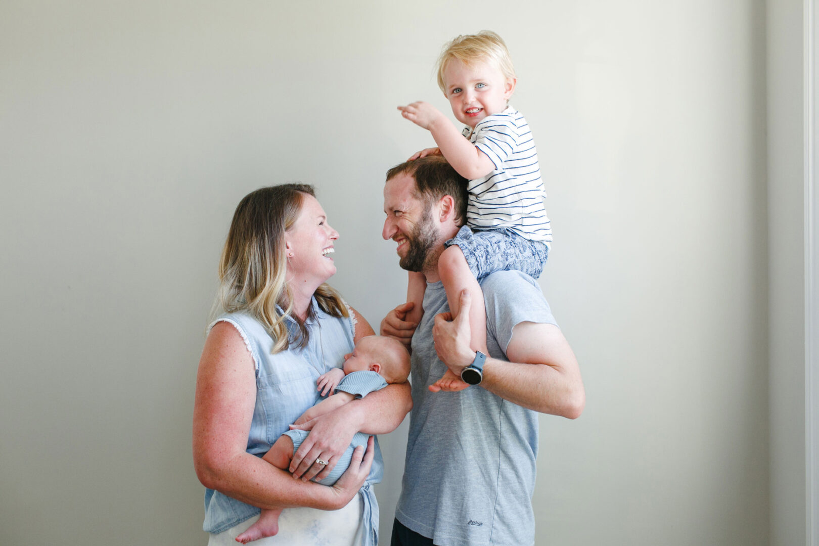 The Collegeville Family Photos beautifully capture a family with two small children; a woman lovingly holds a baby while the man joyfully carries a toddler on his shoulders. They beam happily in front of a light-colored wall, perfectly encapsulating their cherished moments.
