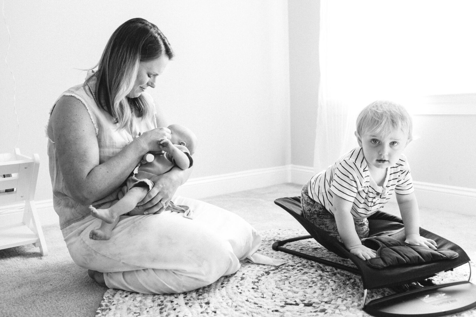 In a sunlit room, a woman sits on the floor bottle-feeding a baby while a young child kneels on a small seat nearby, capturing the warmth of Collegeville family photos.