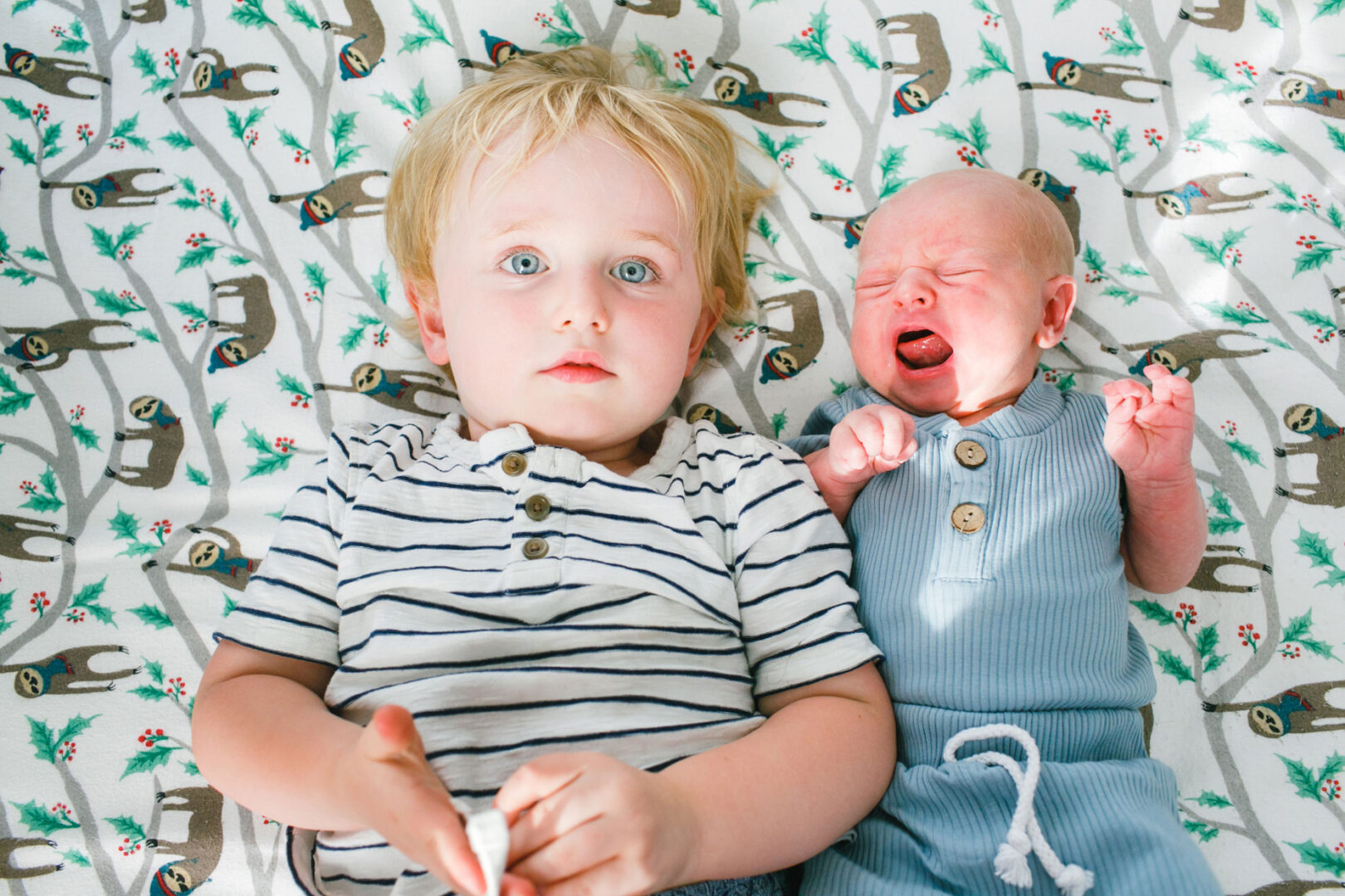 In this Collegeville Family Photos moment, a young boy in a striped shirt lies beside a crying baby on a sloth-patterned blanket. The baby is dressed adorably in blue, capturing the essence of sibling bonds and cozy childhood memories.