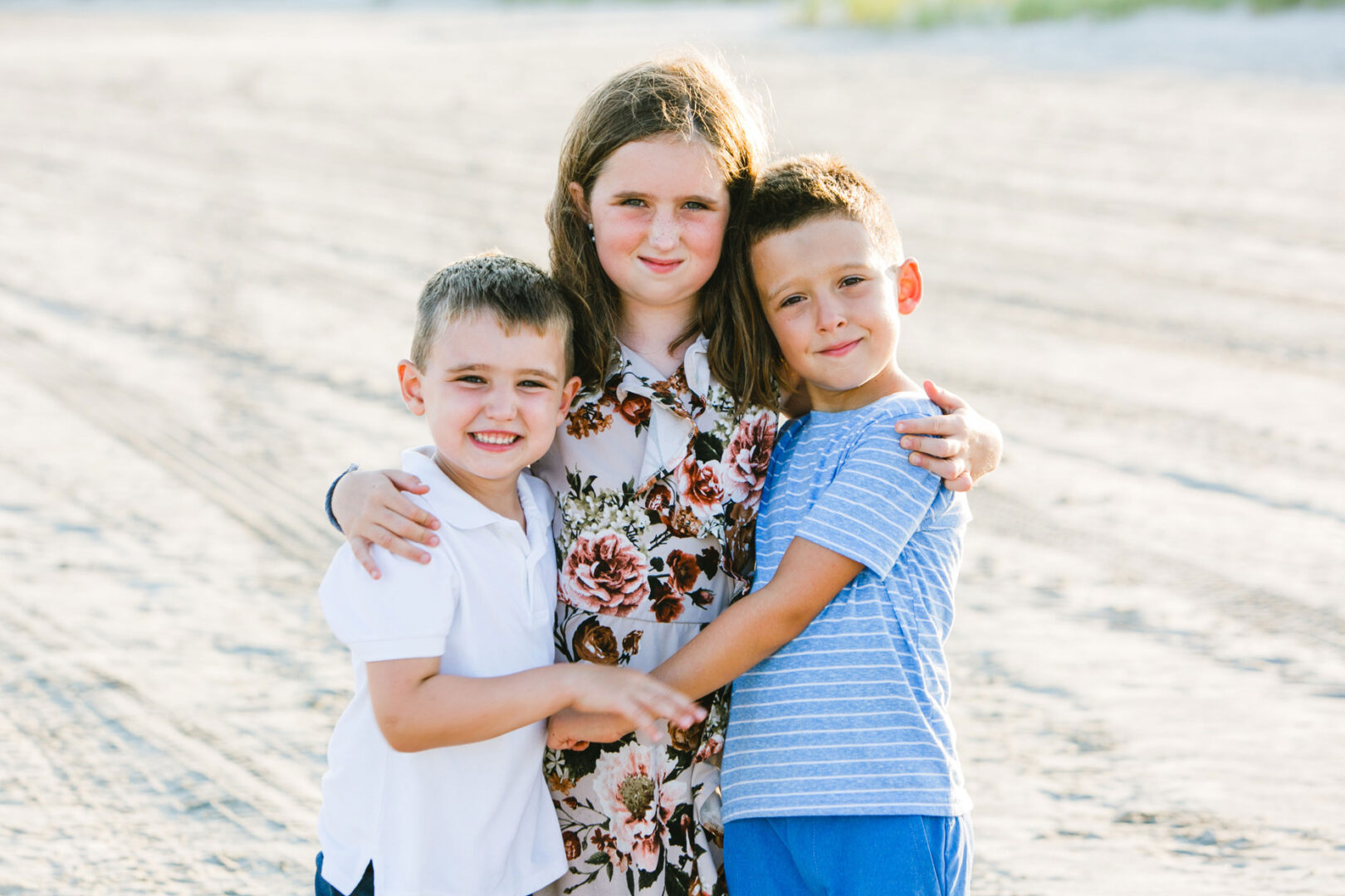Three children stand on a sandy beach, smiling and hugging in a cherished Wildwood family photo. Two boys flank a girl in the center.