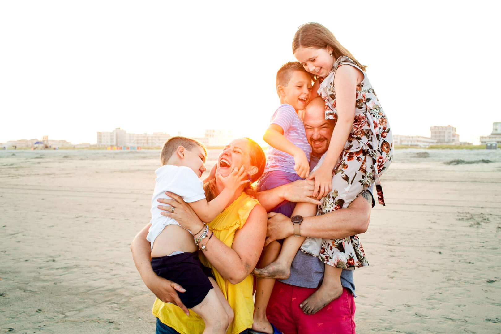 A family of four joyfully laughing and playing on a sandy beach during sunset, capturing perfect Wildwood family photos with buildings silhouetted against the vibrant sky in the background.