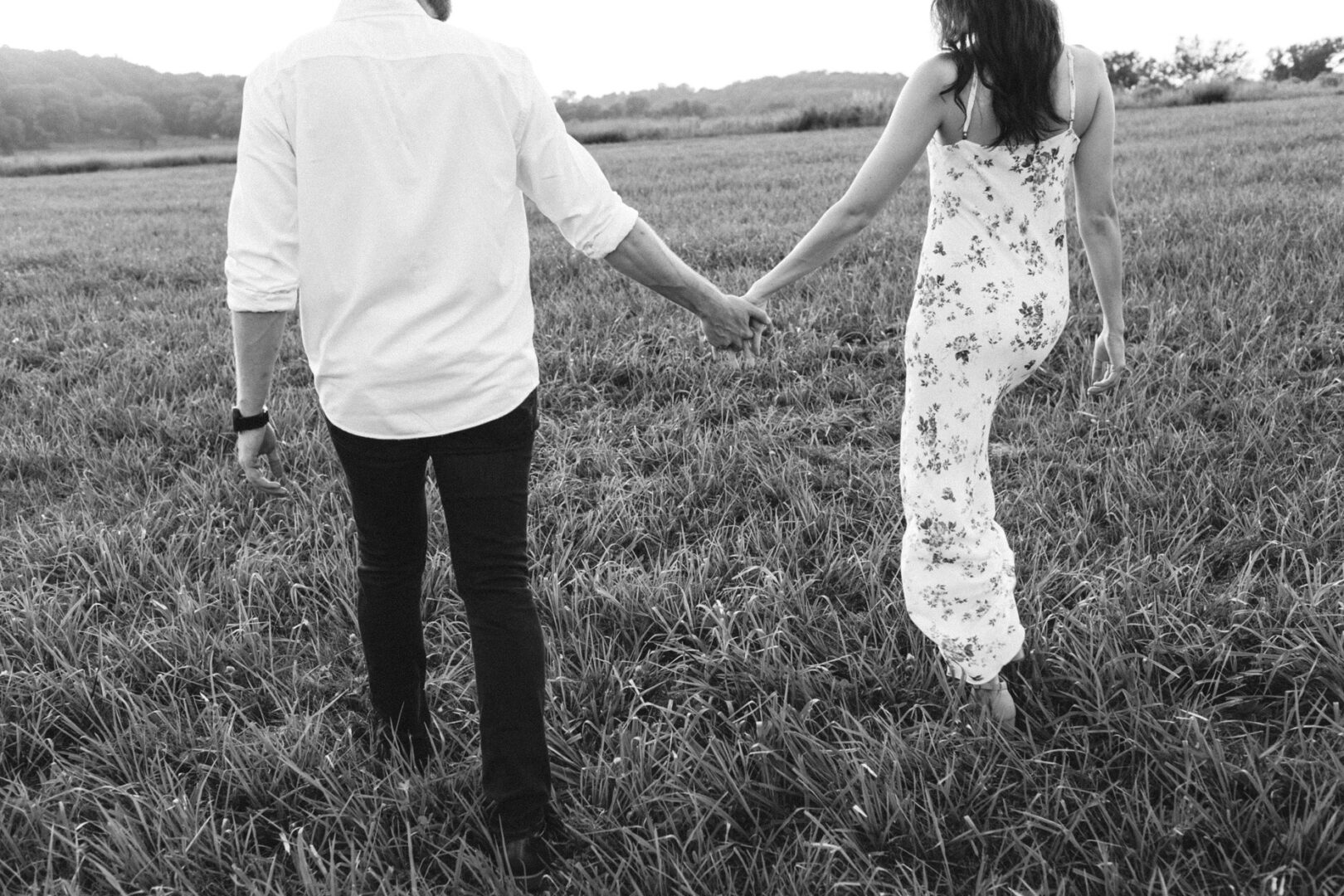 A couple holding hands walks through a grassy field at Valley Forge, both facing away from the camera, capturing the essence of their engagement session.