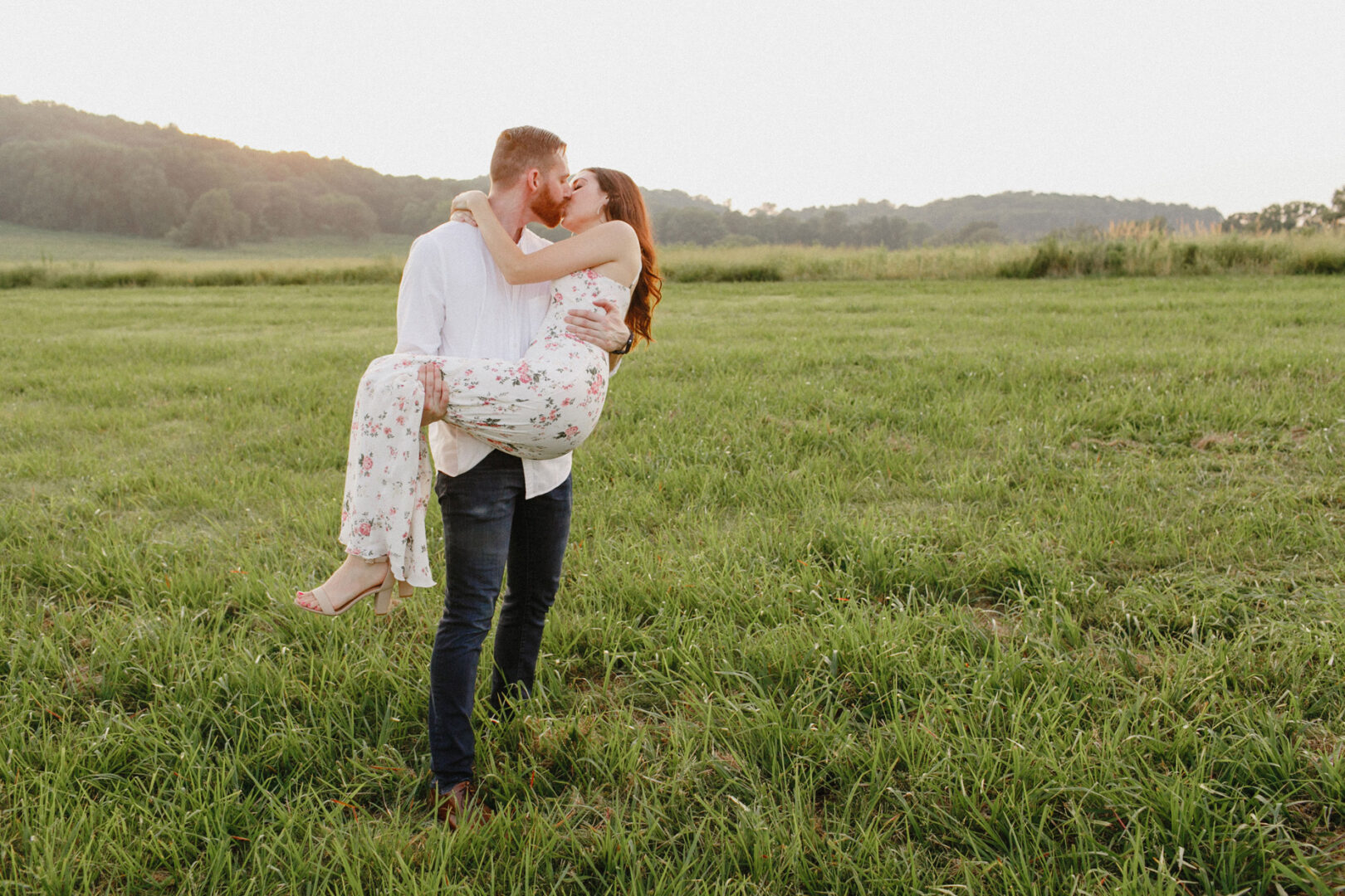 A man in a white shirt and jeans holds a woman in a floral dress, sharing a kiss in the grassy field of Valley Forge during their enchanting engagement session, with rolling hills gracefully framing the background.