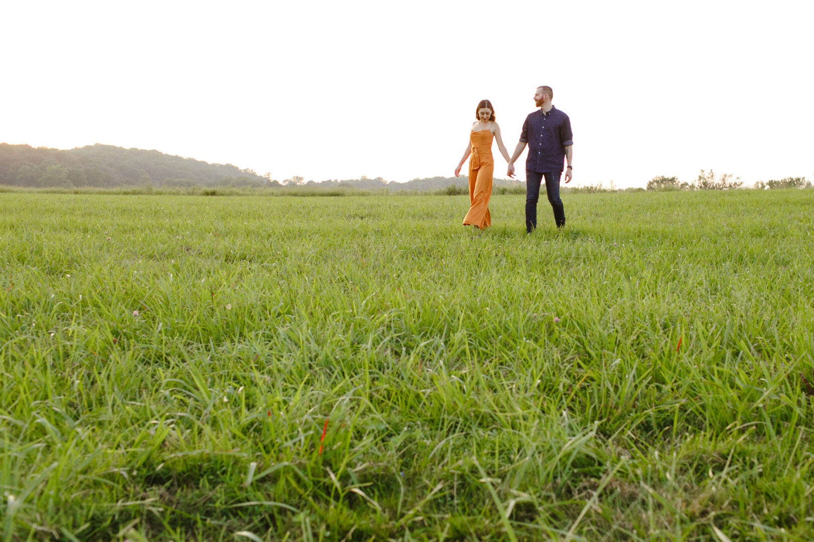 A couple walks hand in hand across a green field under a clear sky, capturing every moment of their Valley Forge engagement session.