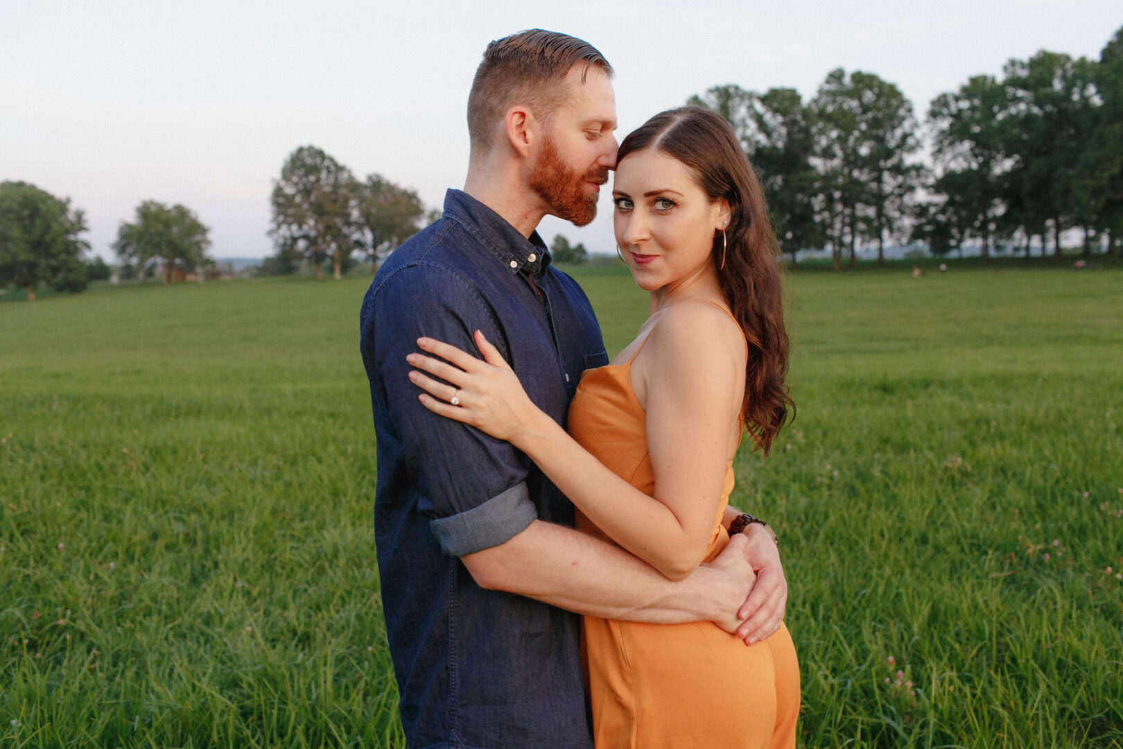 In a beautiful Valley Forge engagement session, a couple embraces in a grassy field. The man, in a blue shirt, gently kisses the woman's forehead as she wears an orange dress and looks at the camera, with trees framing the romantic scene in the background.