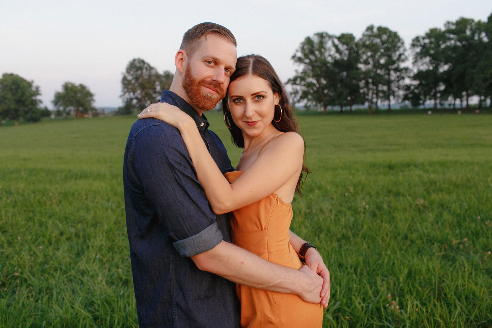 A couple embracing in a grassy field at Valley Forge with trees in the background, both looking at the camera. The woman is wearing an orange dress, and the man is in a blue shirt, capturing a beautiful engagement session.