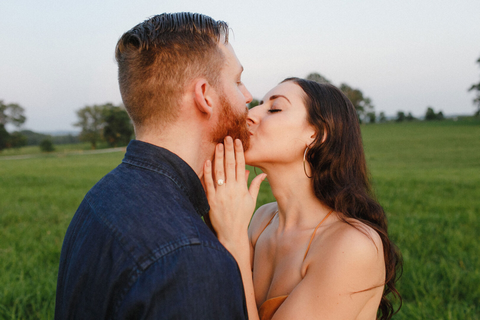 In the midst of a scenic Valley Forge engagement session, a woman kisses a man's cheek in an open field. Her hand, adorned with an engagement ring, rests gently on his face. They are surrounded by grass and trees under the clear sky.