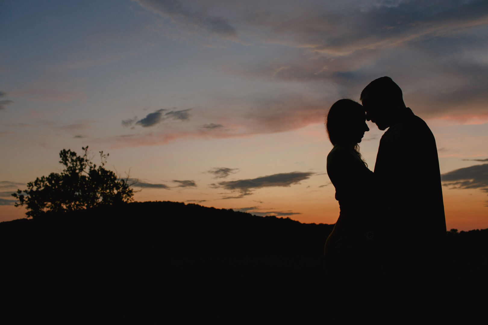 Silhouette of two people facing each other against a sunset sky at Valley Forge, with scattered clouds and a tree in the background, capturing an enchanting engagement session.