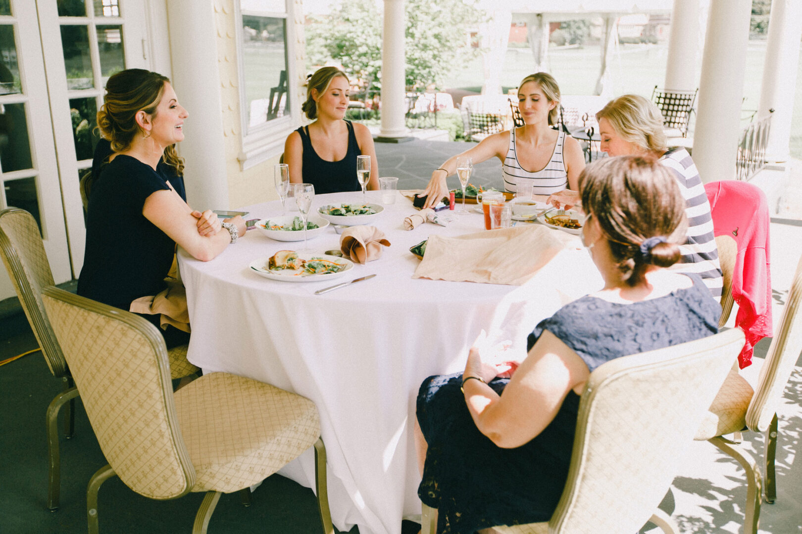 Five women sit around a table sharing a meal on a covered porch, reminiscent of the warm camaraderie often seen at a Philadelphia Cricket Club wedding, with plates of food and beverages in front of them.