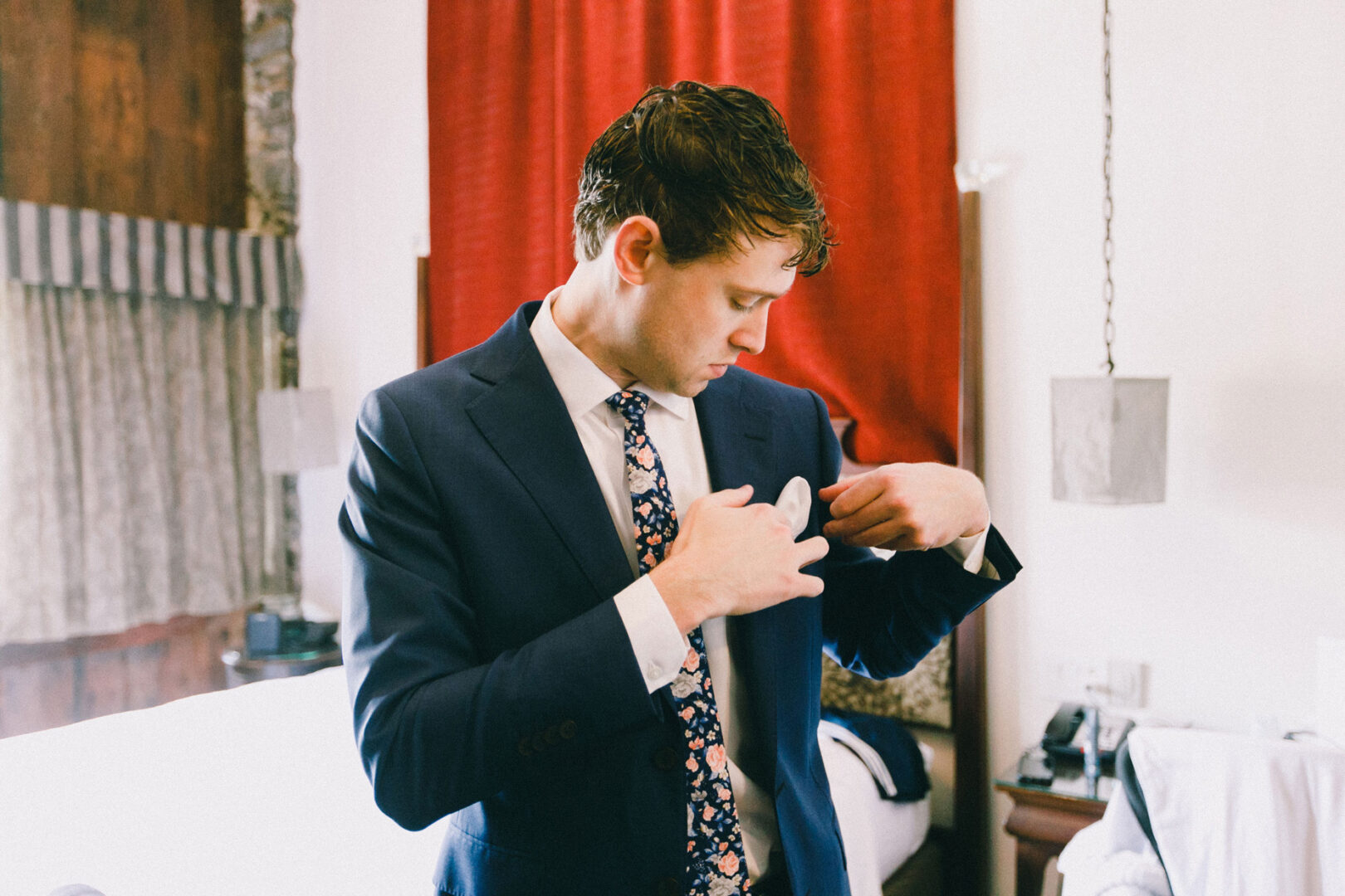 In a hotel room with red curtains and modern decor, a man in a suit adjusts his shirt cuff, preparing for the elegance of a Philadelphia Cricket Club Wedding.