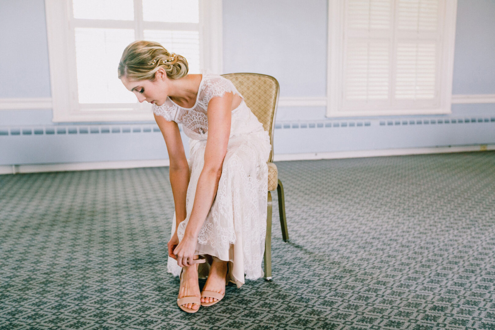 A woman in a lace dress sits on a chair, adjusting her sandal in a sunlit room at the Philadelphia Cricket Club Wedding, with patterned carpet and white shuttered windows enhancing the elegant ambiance.