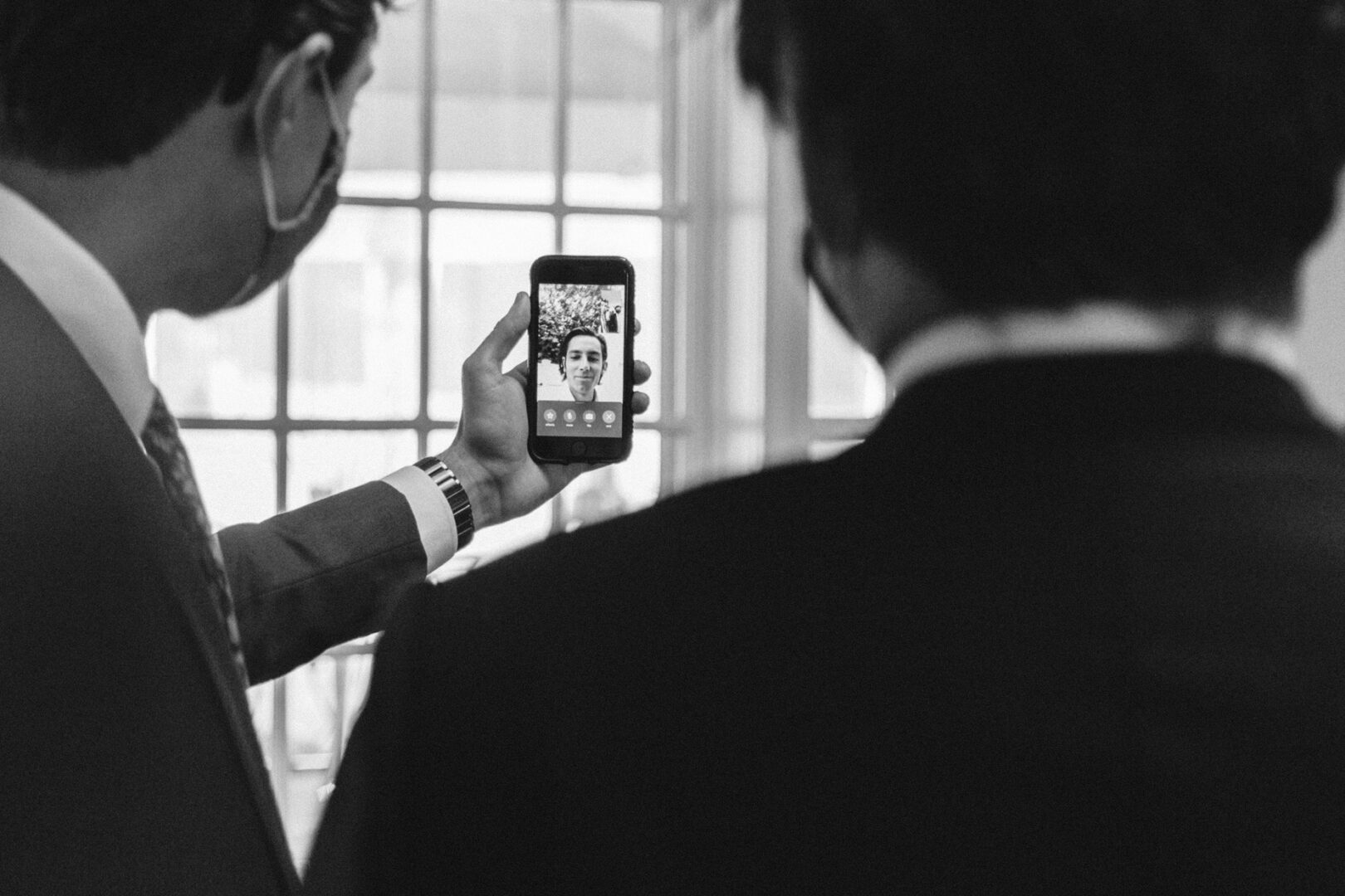Two people in suits look at a phone screen displaying a video call from a lively Philadelphia Cricket Club wedding. One person holds the phone firmly as they share in the celebration from afar. The scene is captured beautifully in black and white.