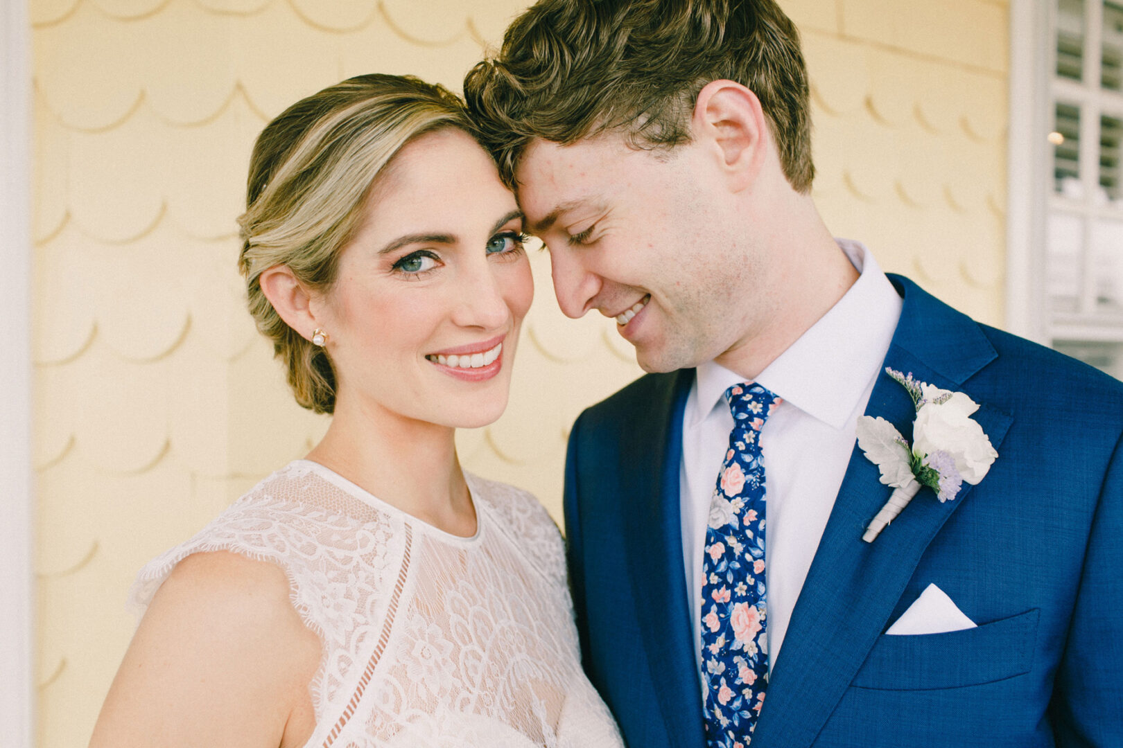 At their Philadelphia Cricket Club wedding, the bride and groom pose together. She wears a lace dress, and he dons a blue suit with a floral tie and boutonniere, standing in front of a yellow wall with scalloped texture.